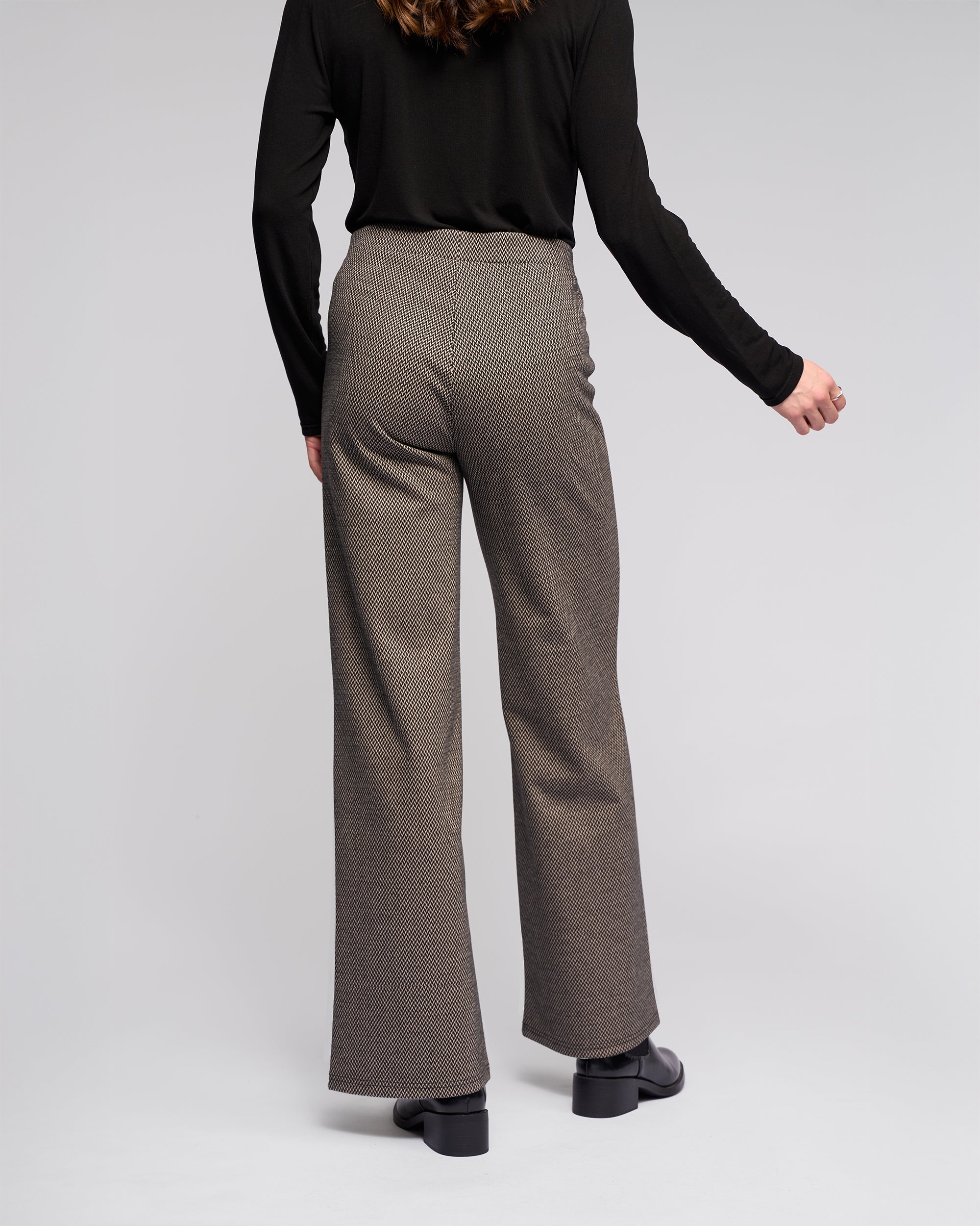 Someone stands facing away, wearing Vassalli Loft Pull On - Knox high-waisted, wide-leg gray stretch knit pants with a black long-sleeve top and black shoes against a plain light background.