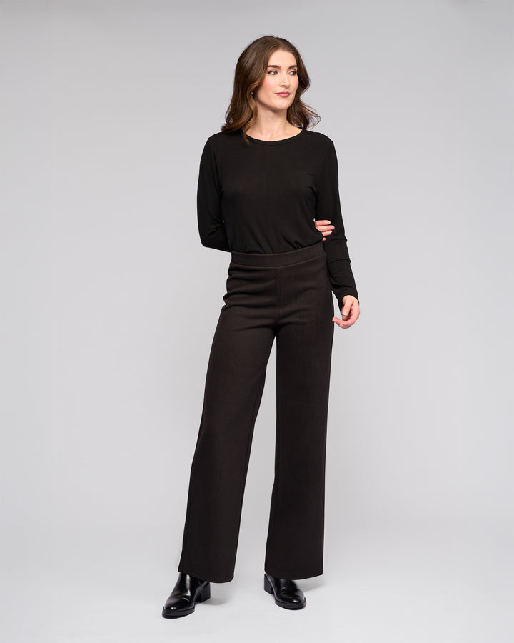 A woman with wavy brown hair wears a long-sleeve black top, black Vassalli Loft Pull On - Arc high-waisted stretch knit wide-leg pants with an elasticated waistband, and black ankle boots, standing against a plain light gray background.