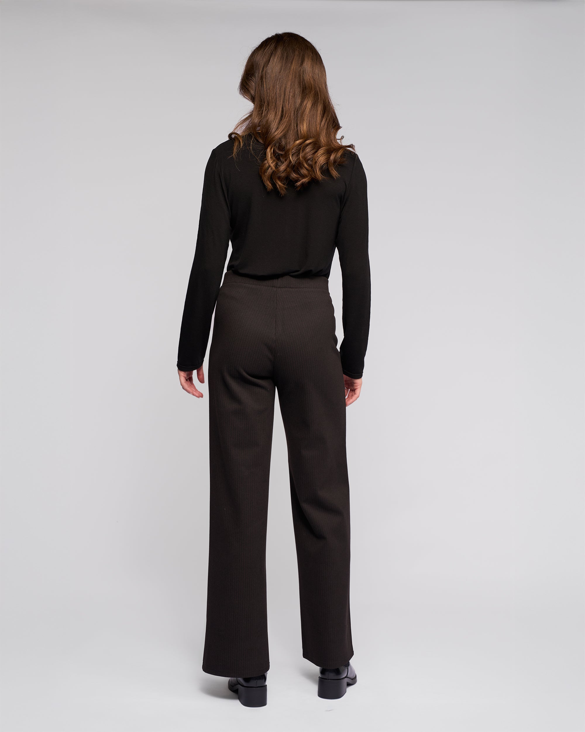 A woman with wavy brown hair stands facing away, wearing Vassalli's Loft Pull On - Metro black long-sleeve top and wide-leg pants with an elasticated waistband, paired with black shoes against a plain light gray background.