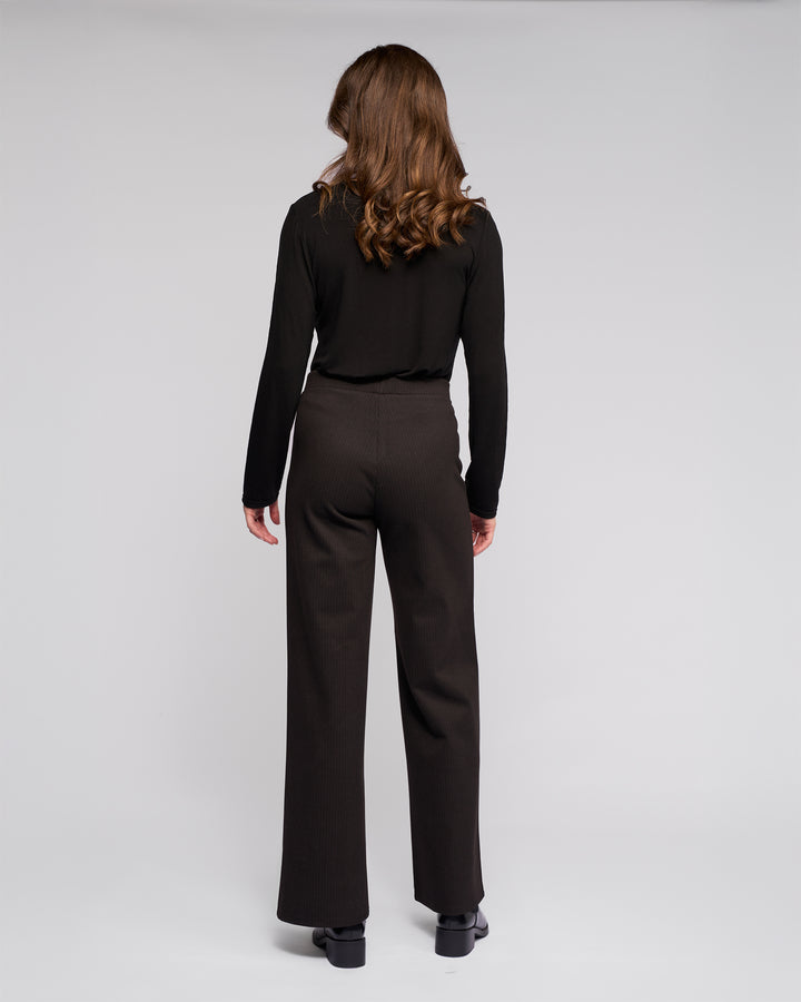 A woman with wavy brown hair stands facing away, wearing Vassalli's Loft Pull On - Metro black long-sleeve top and wide-leg pants with an elasticated waistband, paired with black shoes against a plain light gray background.