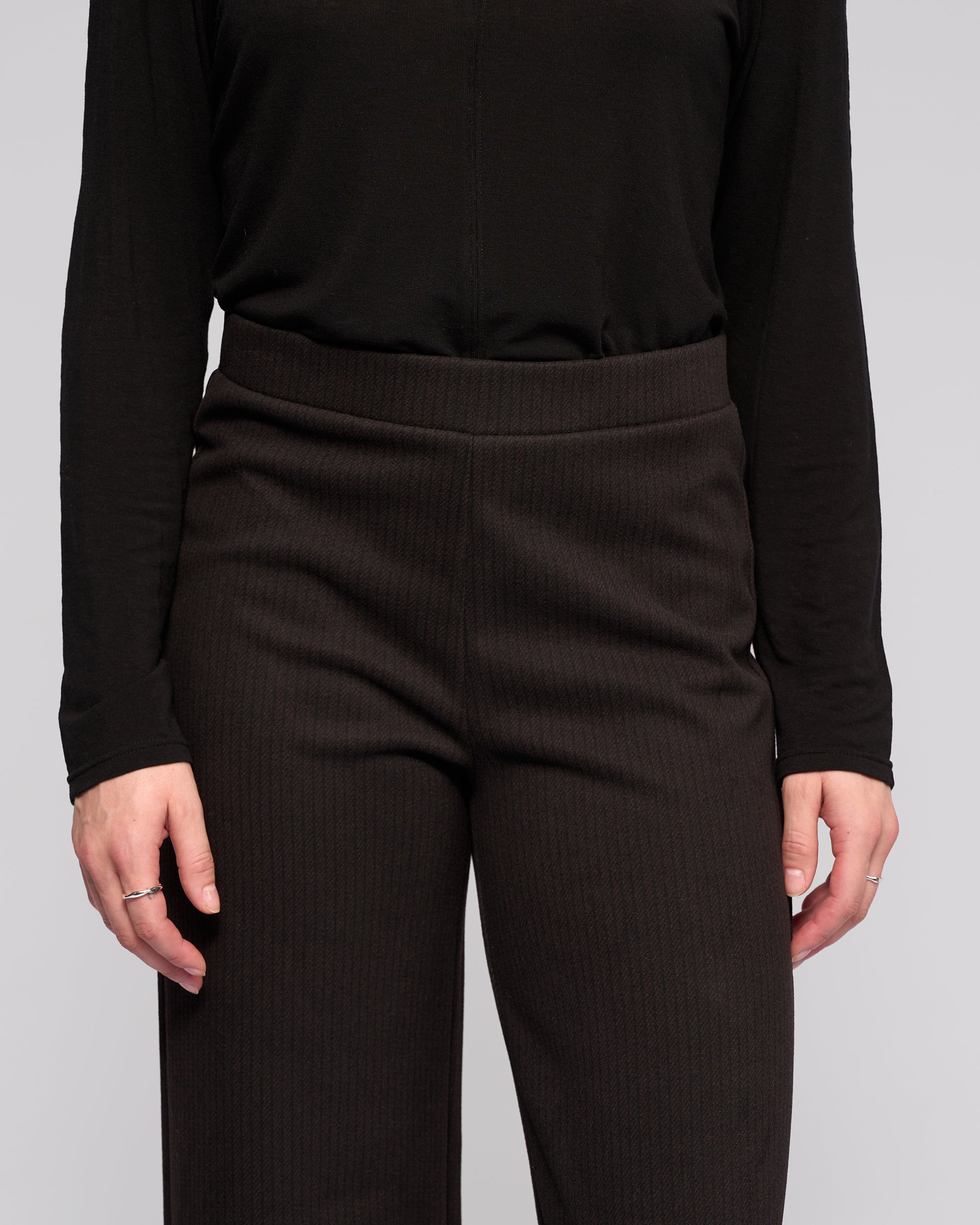 A person wears the Vassalli Loft Pull On - Metro: high-waisted, textured wide-leg pants with an elastic waistband, paired with a long-sleeve black top, standing relaxed with arms at their sides, visible from shoulders to mid-thigh.