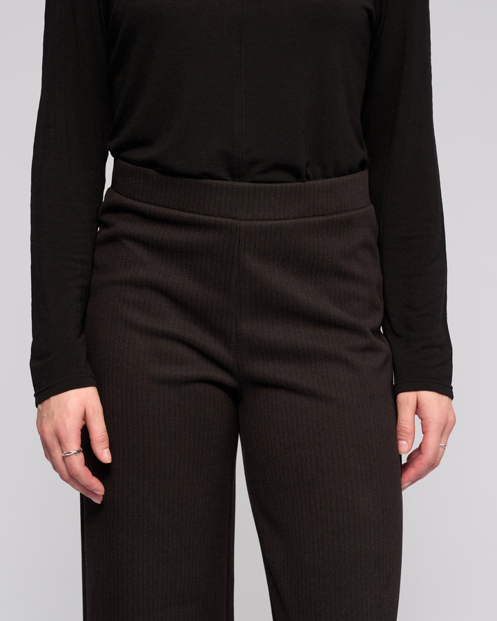 A person wears the Vassalli Loft Pull On - Metro: high-waisted, textured wide-leg pants with an elastic waistband, paired with a long-sleeve black top, standing relaxed with arms at their sides, visible from shoulders to mid-thigh.