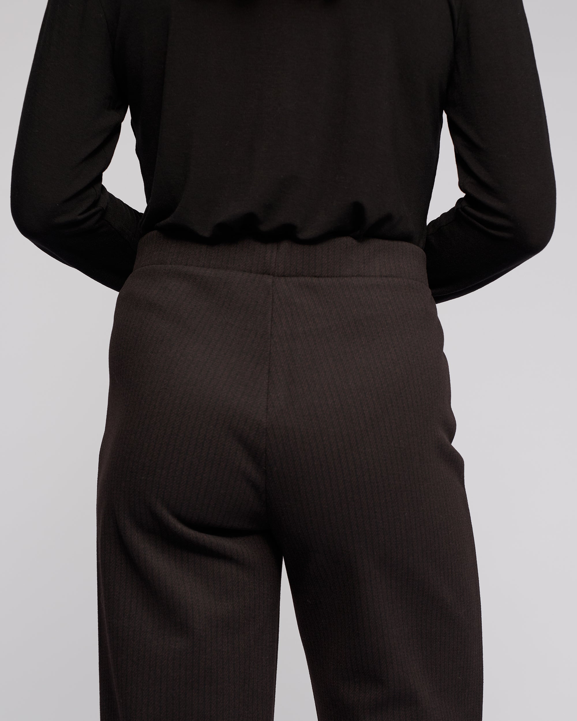 A person wearing Vassalli's Loft Pull On - Metro stands with their arms behind their back, facing away from the camera. The outfit features a black long-sleeve top and stretch knit wide-leg pants.