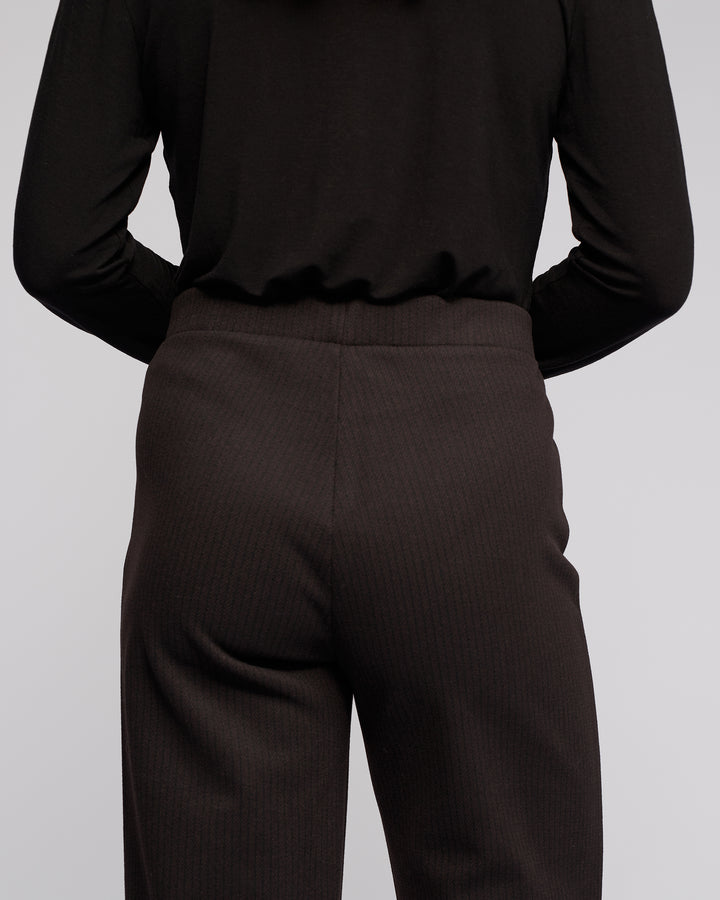 A person wearing Vassalli's Loft Pull On - Metro stands with their arms behind their back, facing away from the camera. The outfit features a black long-sleeve top and stretch knit wide-leg pants.