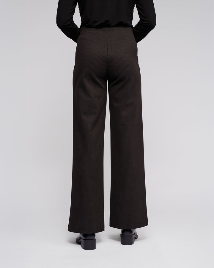A person wearing Vassalli Loft Pull On - Metro black, high-waisted, wide-leg pants with an elasticated waistband and black shoes stands facing away from the camera against a plain light background.