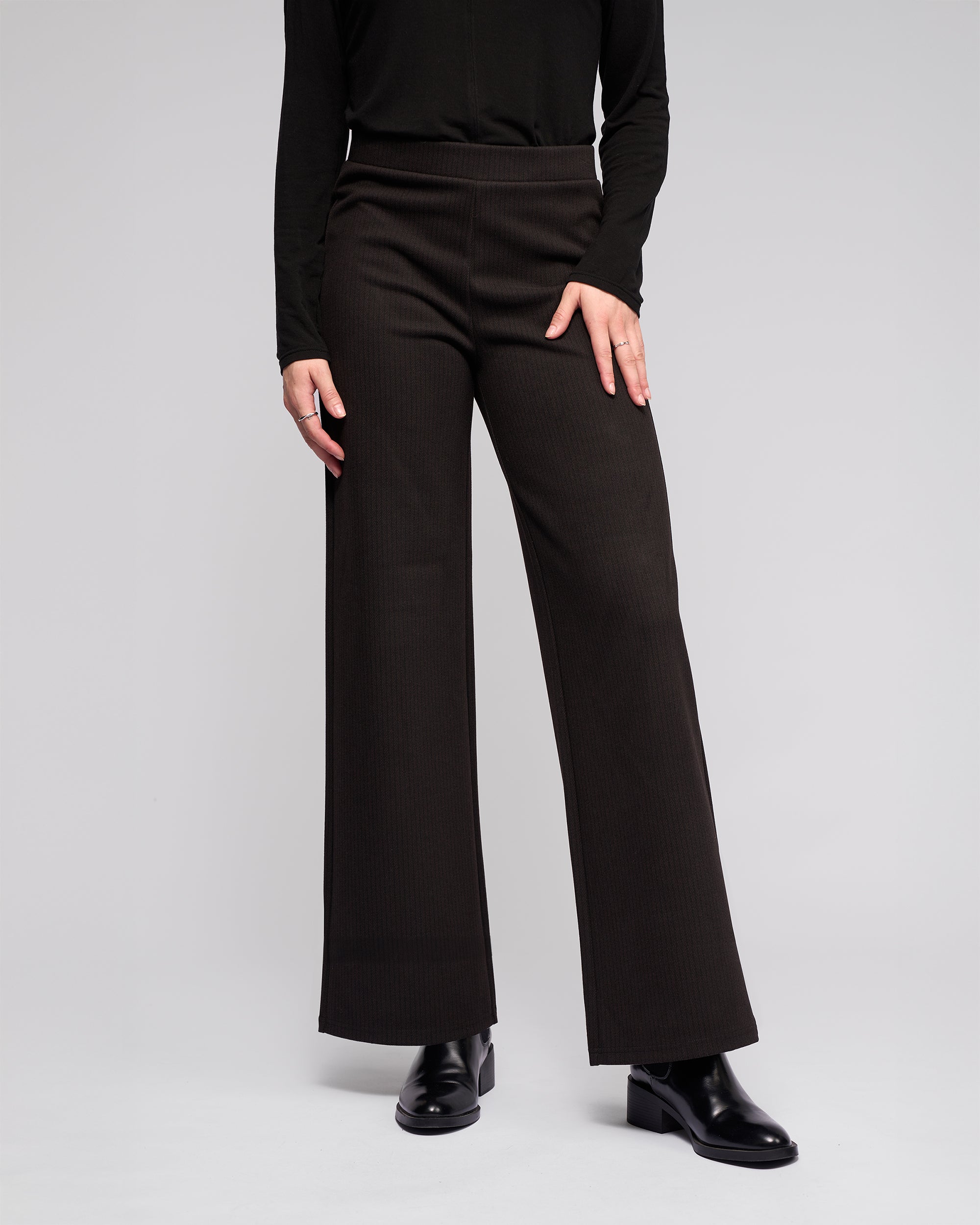 A person wearing the Vassalli Loft Pull On - Metro black wide-leg pants, a black long-sleeve top, and black boots stands against a plain light gray background. Their upper face is not visible in the image.