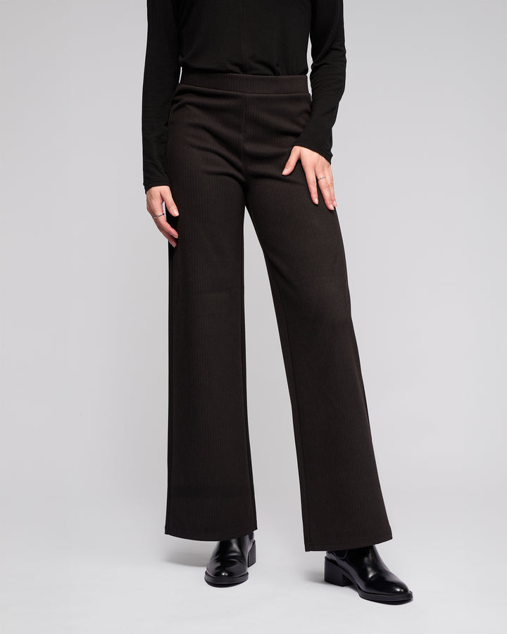 A person wearing the Vassalli Loft Pull On - Metro black wide-leg pants, a black long-sleeve top, and black boots stands against a plain light gray background. Their upper face is not visible in the image.