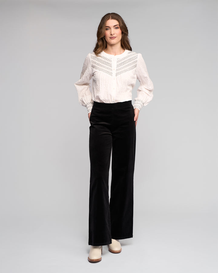 A woman stands against a light background wearing a white lace-detailed blouse and Vassalli Loft Cord Pull On pants in black, featuring a wide-leg design and elastic waistband. She faces forward with her hands in the pockets, paired with light heeled boots.