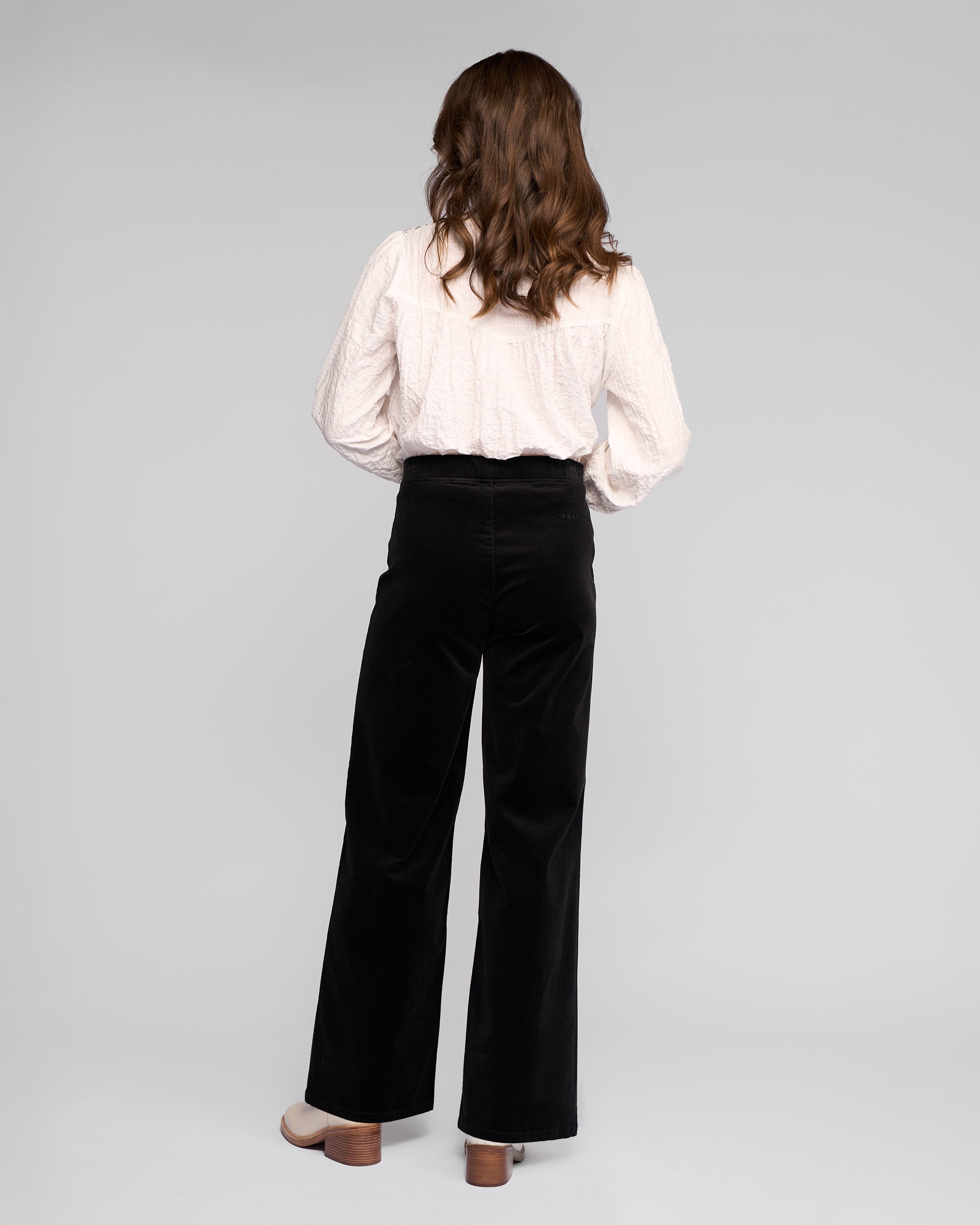 A woman with wavy brown hair faces away, wearing a textured white long-sleeve blouse and Vassalli Loft Cord Pull On - Black wide-leg pants with an elasticated waistband, plus beige heeled shoes, against a plain light gray background.