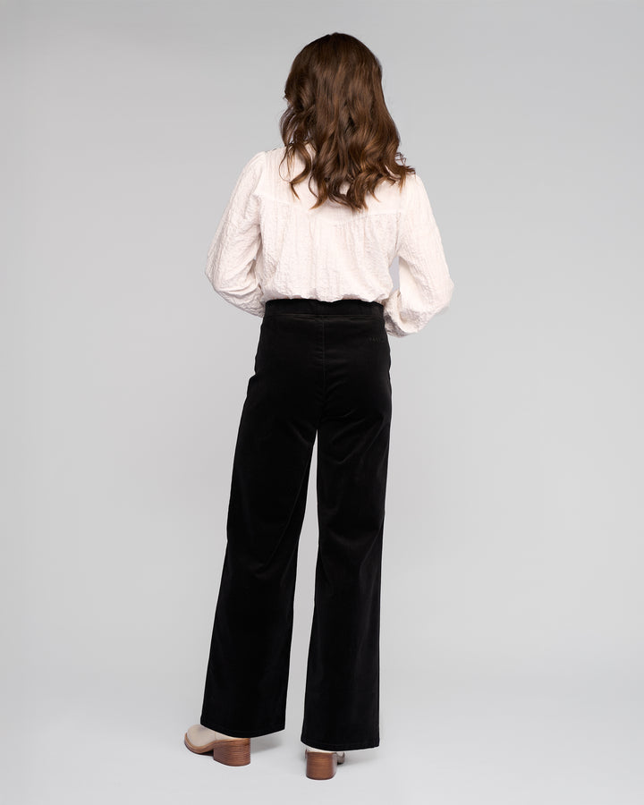 A woman with wavy brown hair faces away, wearing a textured white long-sleeve blouse and Vassalli Loft Cord Pull On - Black wide-leg pants with an elasticated waistband, plus beige heeled shoes, against a plain light gray background.