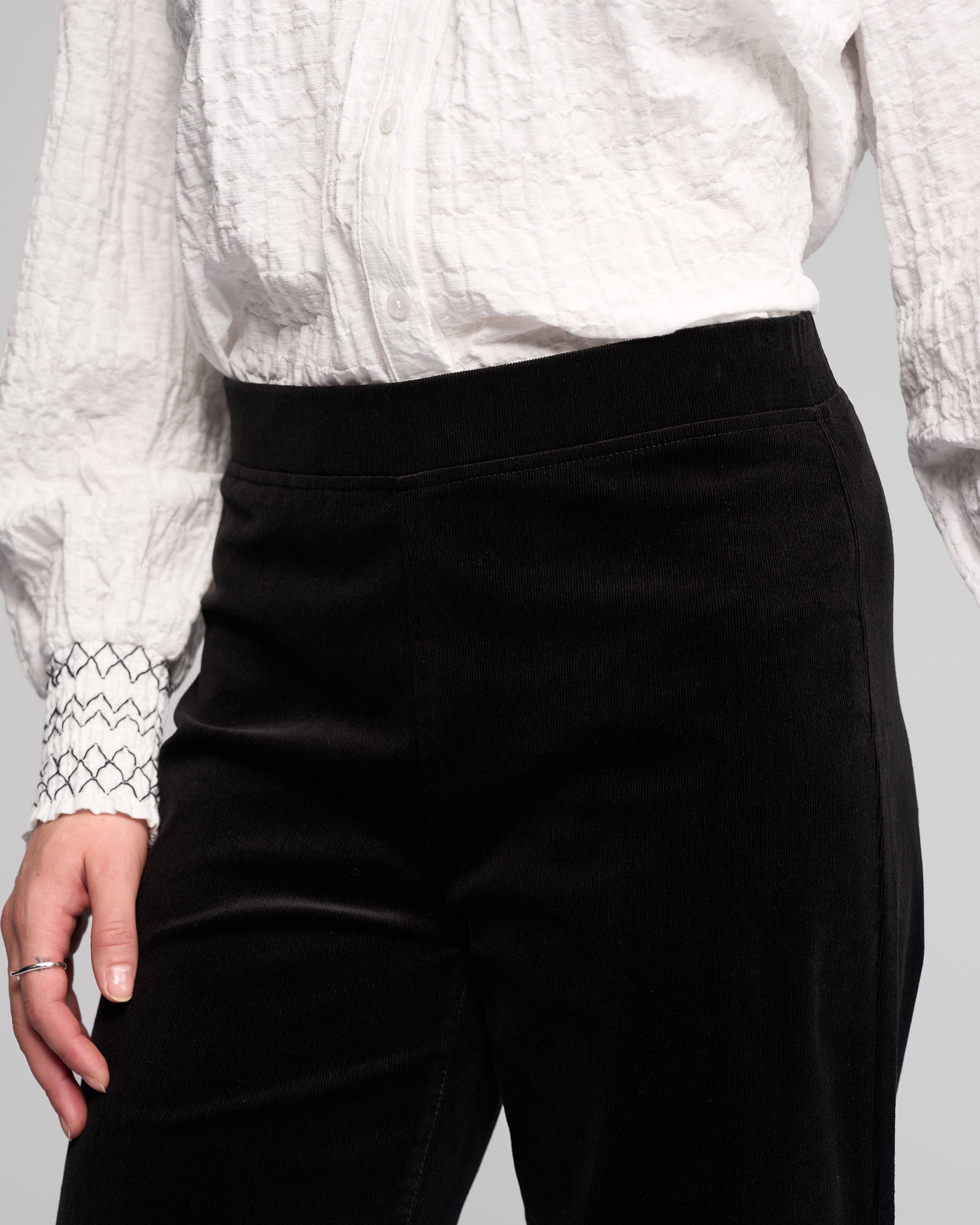 A person wearing black Vassalli Loft Cord Pull On pants and a white textured button-up shirt with patterned cuffs, shown from the chest down with one hand by their side.