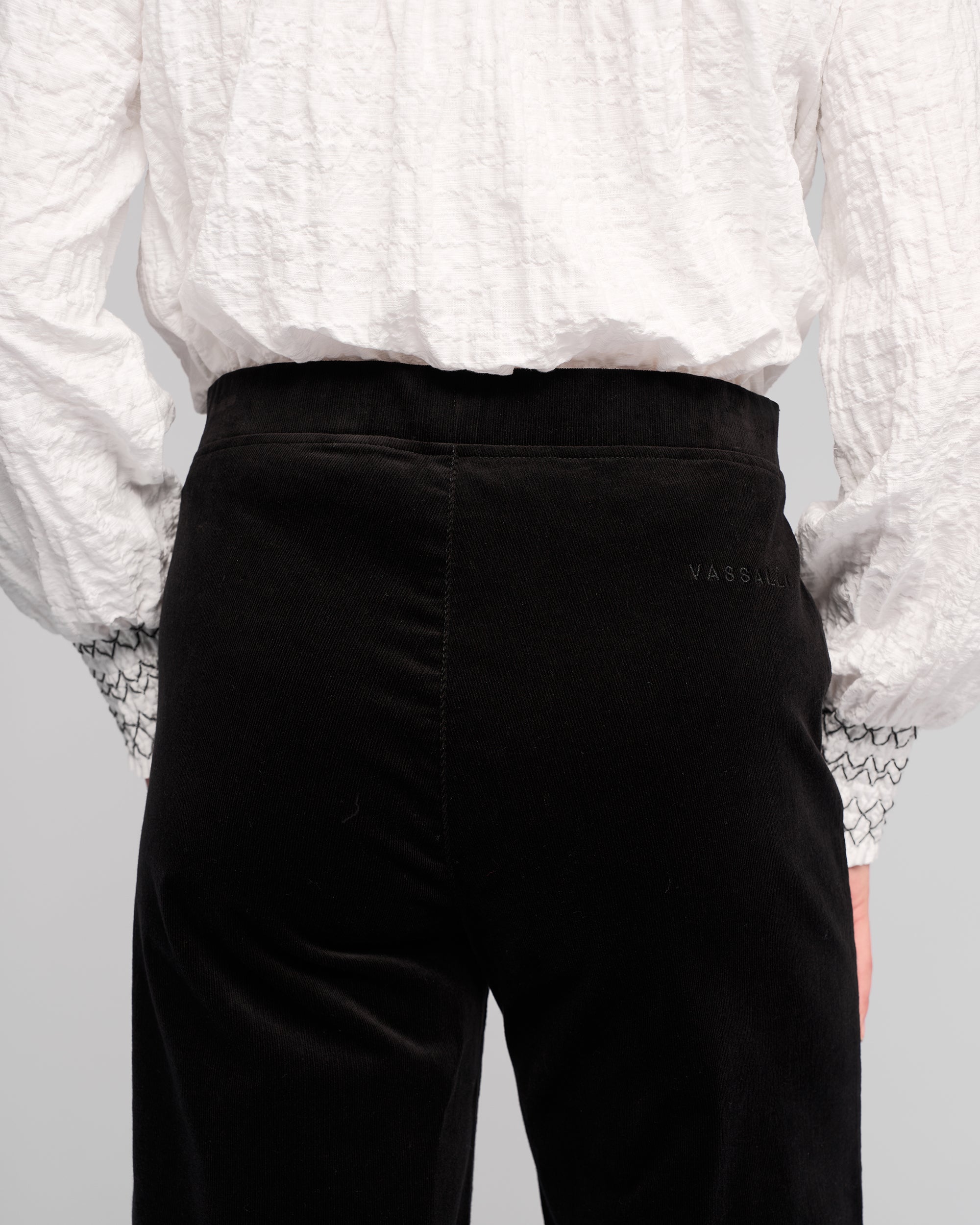 Model wears a textured white long-sleeve shirt with patterned cuffs and Vassalli Loft Cord Pull On - Black pinwale cord wide-leg pants, shown from the back. Subtle text detail is visible near the waistband on the right side of the pants.
