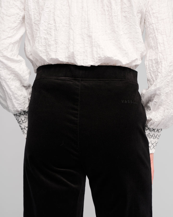 Model wears a textured white long-sleeve shirt with patterned cuffs and Vassalli Loft Cord Pull On - Black pinwale cord wide-leg pants, shown from the back. Subtle text detail is visible near the waistband on the right side of the pants.