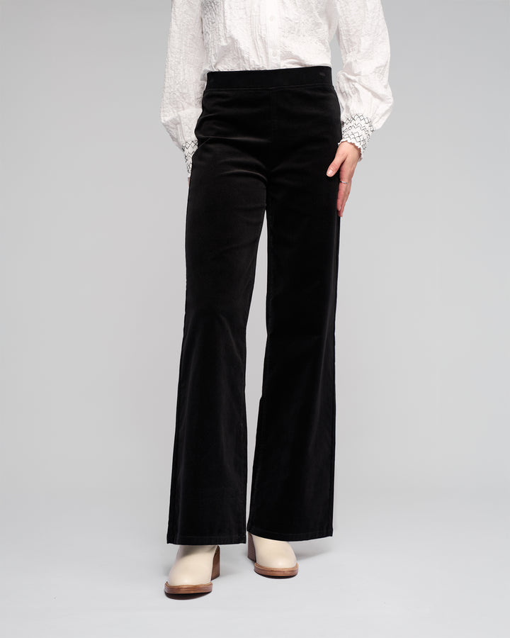 Model wears the Vassalli Loft Cord Pull On pants in black with a white textured long-sleeve blouse and cream heeled boots, standing against a plain light gray background.