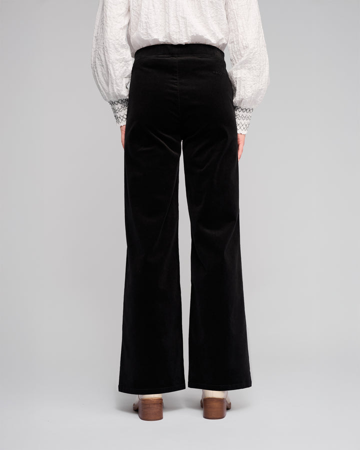 A person stands facing away, wearing Vassalli's Loft Cord Pull On - Black pants with an elasticated waistband, a white textured blouse with patterned cuffs, and brown heeled shoes against a plain light background.