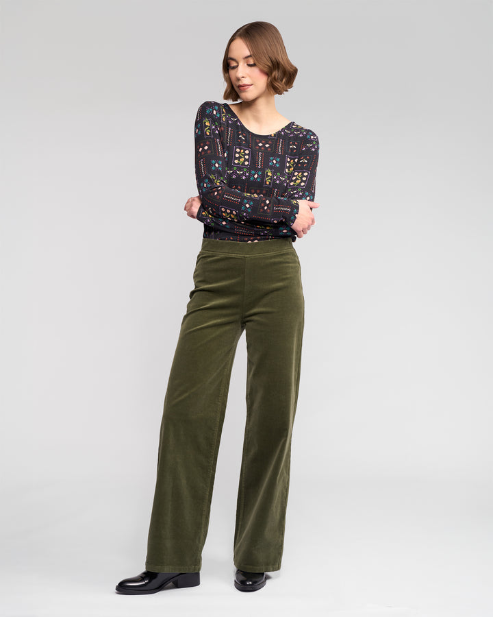 A woman stands against a plain background, wearing a patterned long-sleeve top, black shoes, and Vassalli’s Loft Cord Pull On wide-leg pants in Deep Sage. She looks slightly down with her arms crossed.