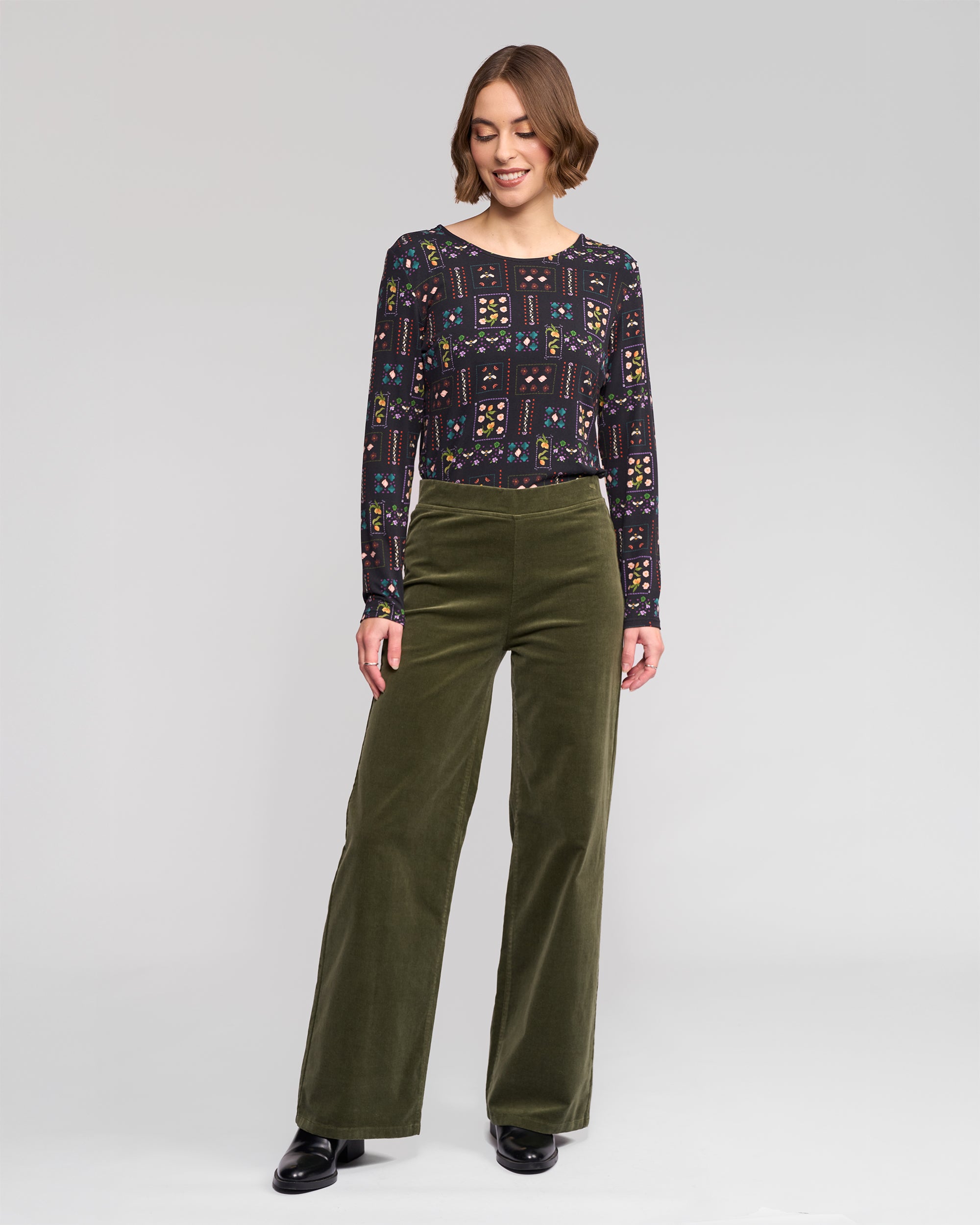 A woman smiles in a patterned dark long-sleeve top and Vassalli Loft Cord Pull On - Deep Sage wide-leg pants, paired with black shoes, against a plain light background.