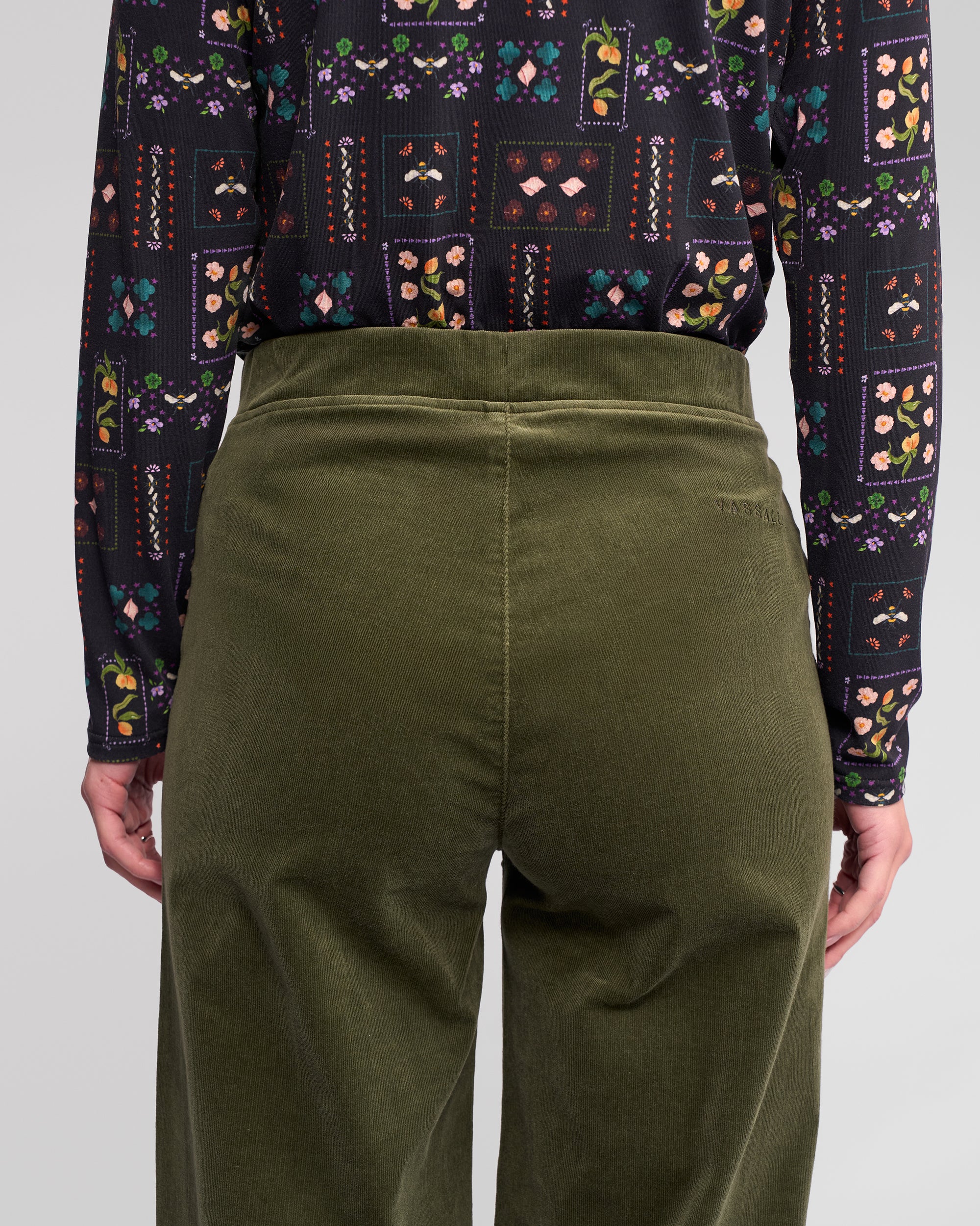 A person shown from the back, waist to mid-thigh, is wearing Vassalli's Loft Cord Pull On pants in Deep Sage and a long-sleeve black shirt with a colorful floral and geometric pattern.