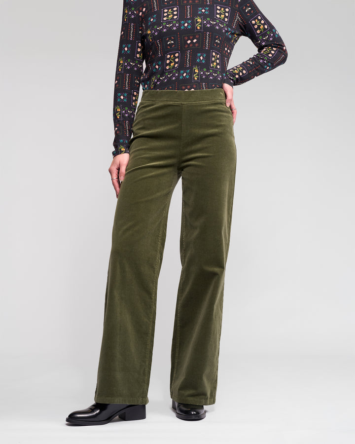 A person wears Vassalli's Loft Cord Pull On pants in deep sage, a patterned long-sleeve top, and black shoes, standing with one hand on their hip against a plain light gray background.