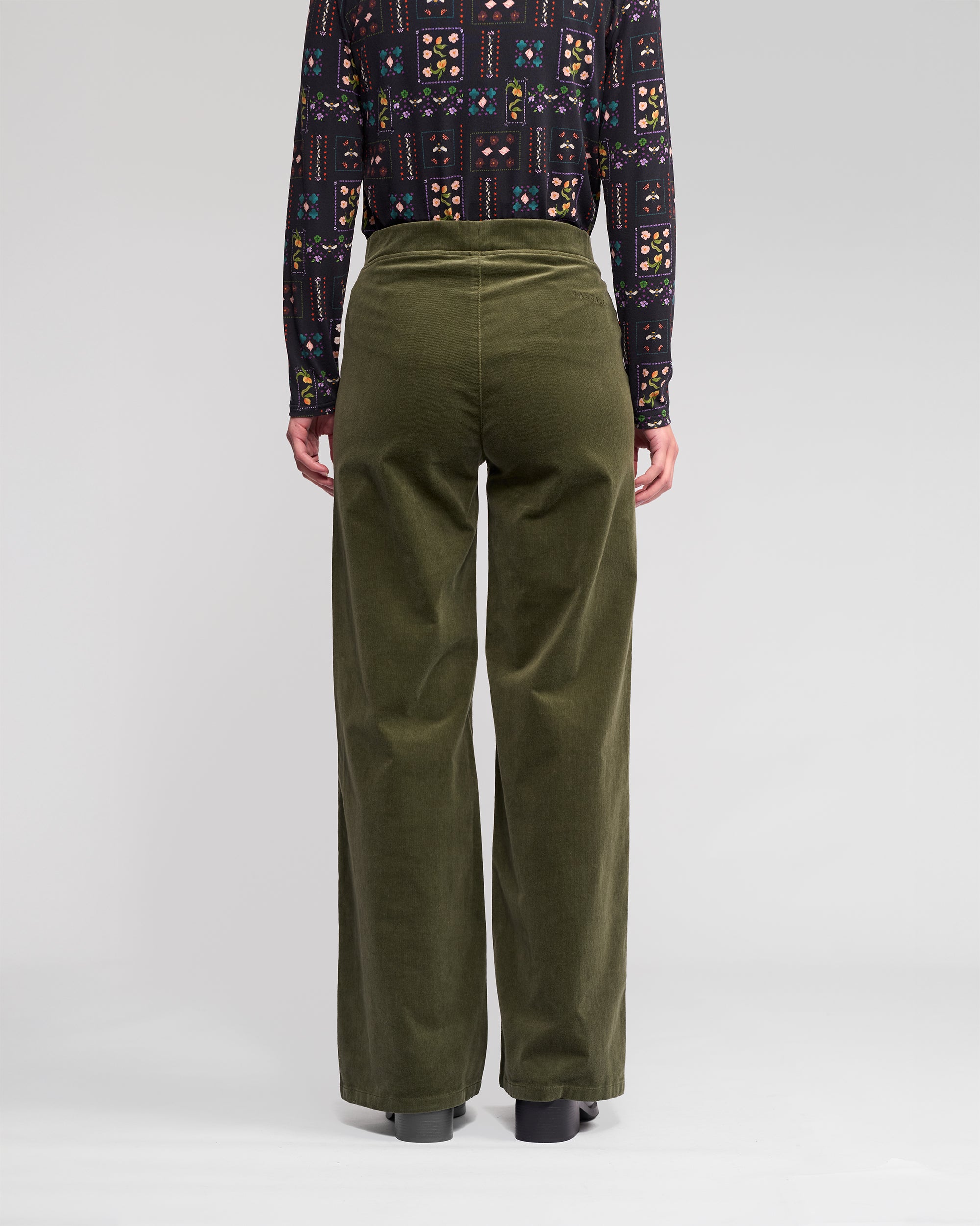 A person faces away, wearing Vassalli Loft Cord Pull On pants in Deep Sage with a dark patterned long-sleeve shirt and black shoes, standing on a plain light background.
