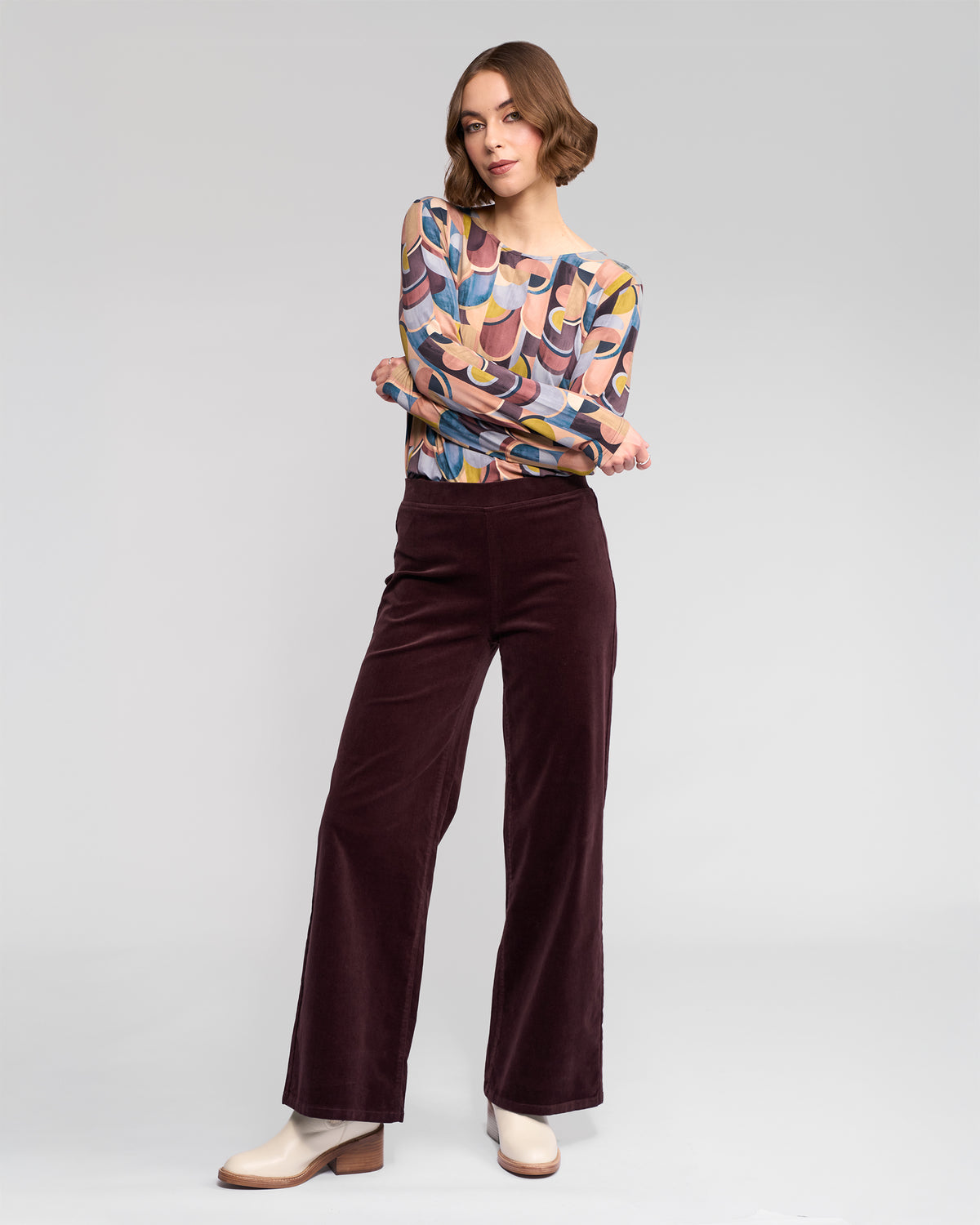 A woman poses with arms crossed and a slight smile against a light gray background, wearing the Vassalli Loft Cord Pull On pant in Merlot, an abstract-patterned long-sleeve top, and cream ankle boots.
