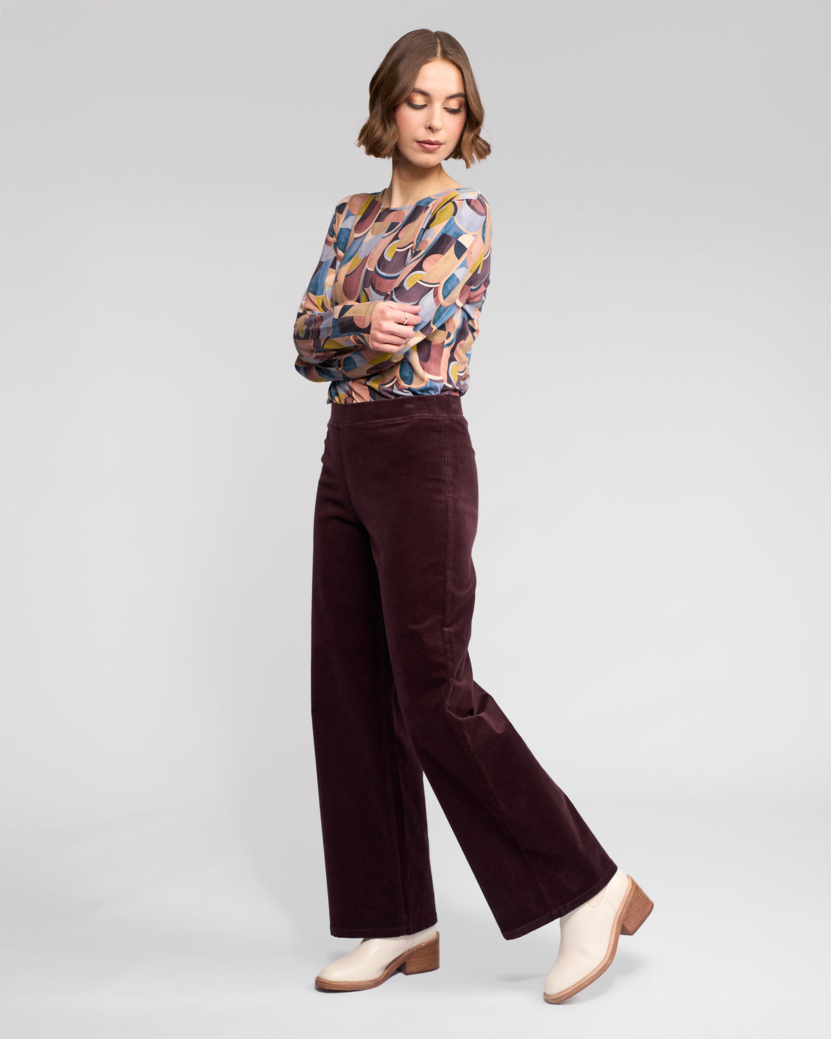 A woman with short brown hair poses in a colorful patterned long-sleeve top, Vassalli Loft Cord Pull On - Merlot wide-leg pants, and white ankle boots against a plain light background.