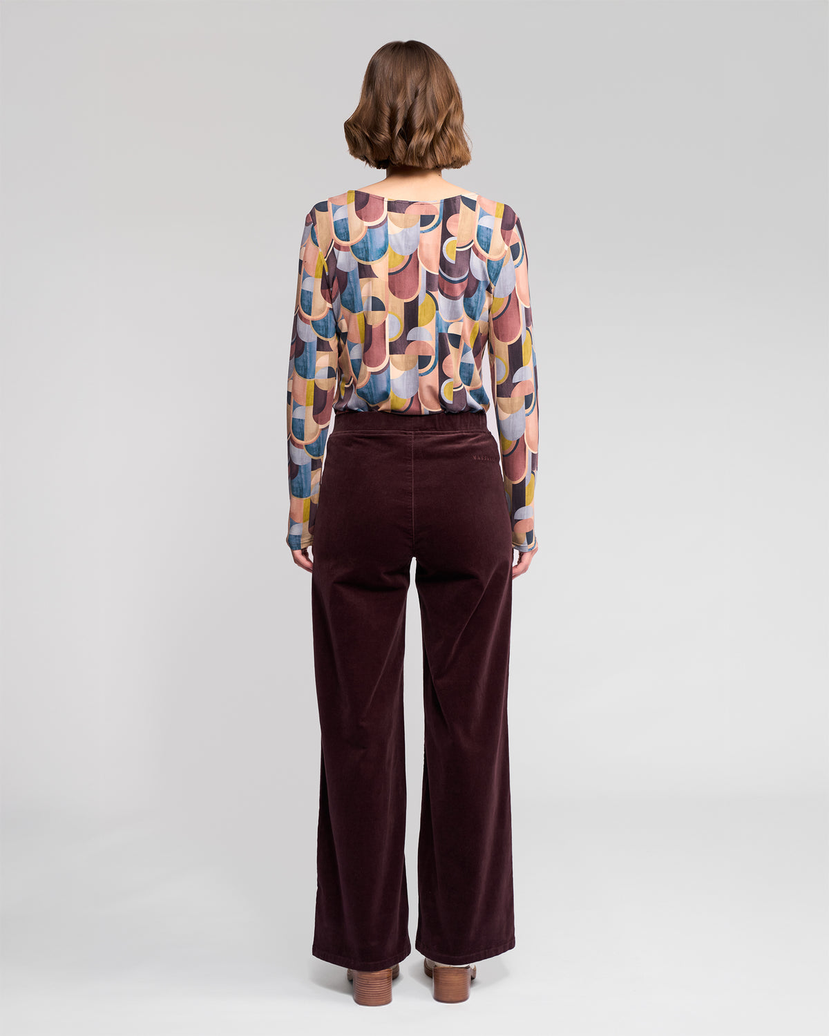 A woman with short brown hair, standing against a light gray background, models the Vassalli Loft Cord Pull On pants in Merlot—a high-waisted, wide-leg pinwale cord style—paired with a colorful geometric long-sleeve top.