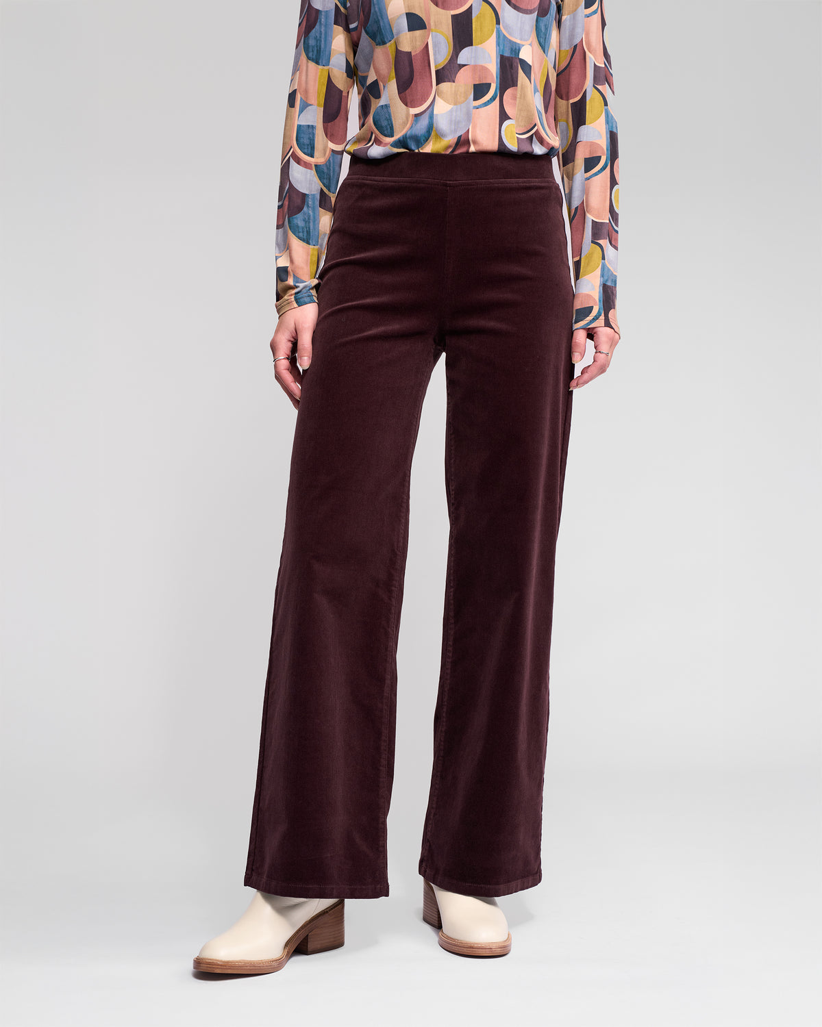 A person wears Vassalli Loft Cord Pull On pants in Merlot with a colorful geometric long-sleeve blouse and beige heeled ankle boots, standing against a plain light background.