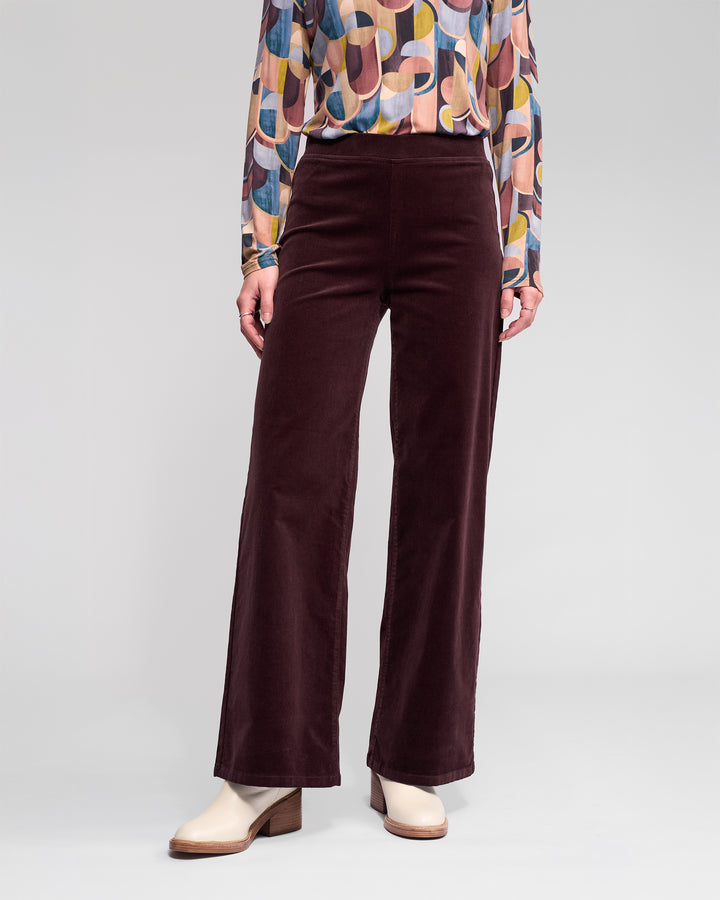 A person wears Vassalli Loft Cord Pull On pants in Merlot with a colorful geometric long-sleeve blouse and beige heeled ankle boots, standing against a plain light background.