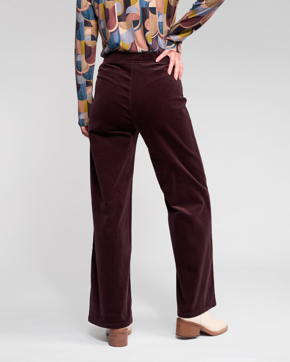 A person in Vassalli's Loft Cord Pull On pants in Merlot, a vibrant geometric long-sleeve shirt, and beige heeled shoes stands with one hand on their waist, seen from the back.