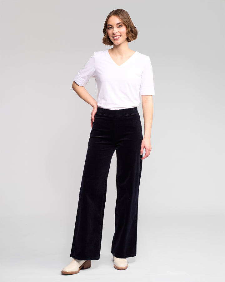 A woman with short, wavy brown hair smiles against a light background, wearing a white V-neck shirt, Vassalli Loft Cord Pull On - Midnight high-waisted wide-leg pants, and light heeled shoes.