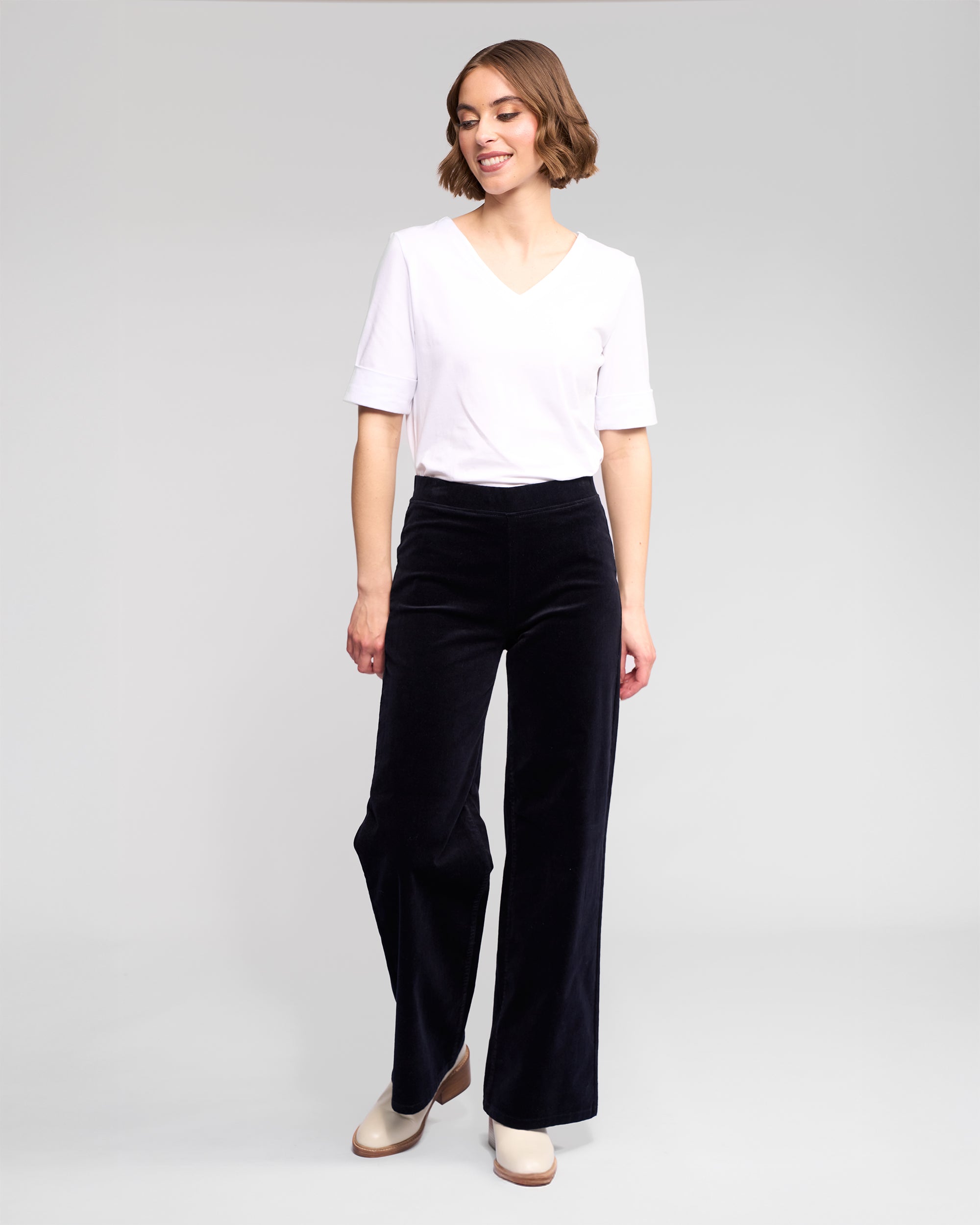 A woman smiles, wearing a white short-sleeve V-neck top, beige shoes, and Vassalli Loft Cord Pull On wide-leg pants in Midnight, posing against a plain light gray background.