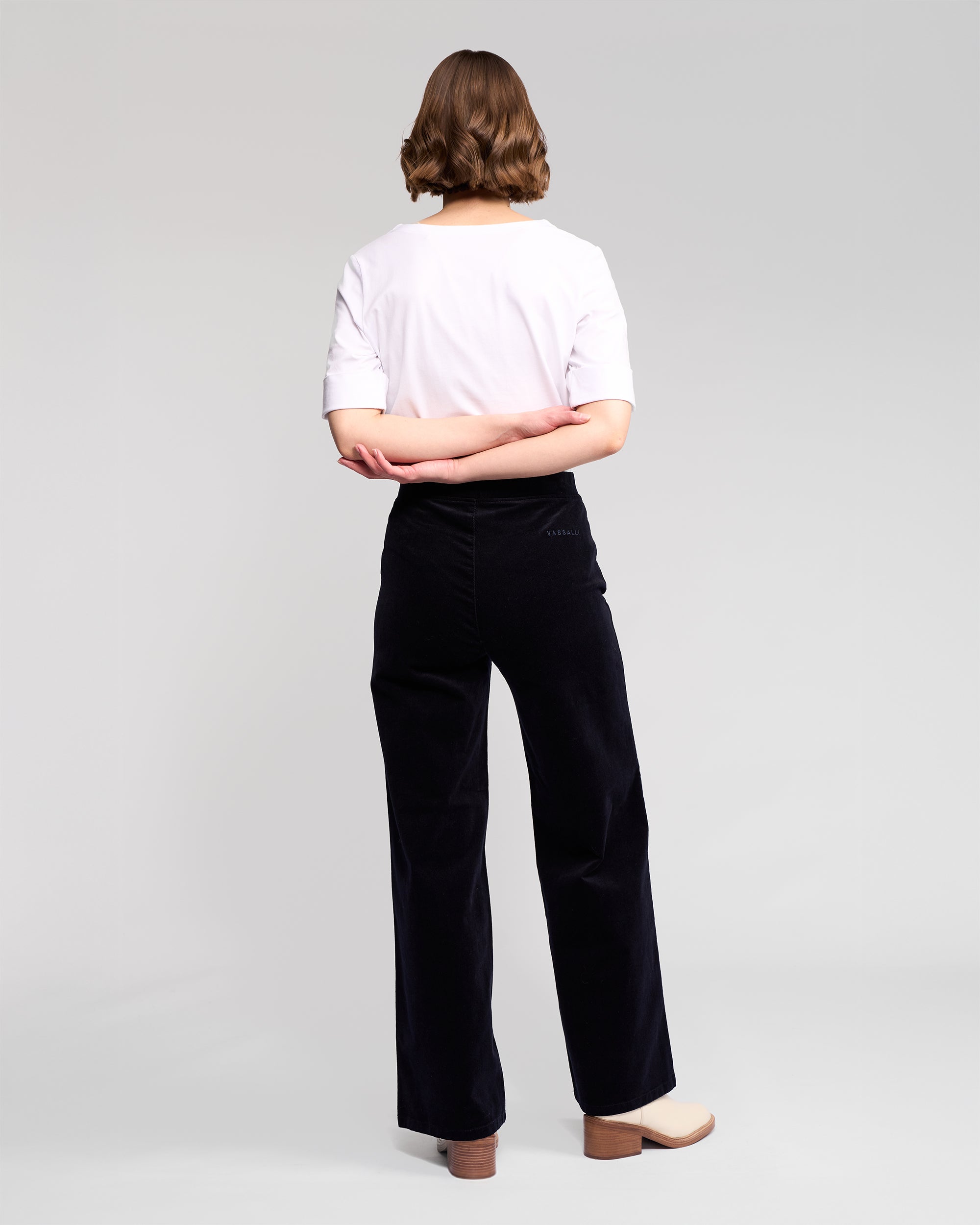 A person with short, wavy brown hair stands with their back to the camera, wearing a white t-shirt and Vassalli Loft Cord Pull On - Midnight pants, paired with beige platform shoes, against a plain light gray background.