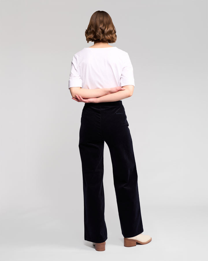 A person with short, wavy brown hair stands with their back to the camera, wearing a white t-shirt and Vassalli Loft Cord Pull On - Midnight pants, paired with beige platform shoes, against a plain light gray background.