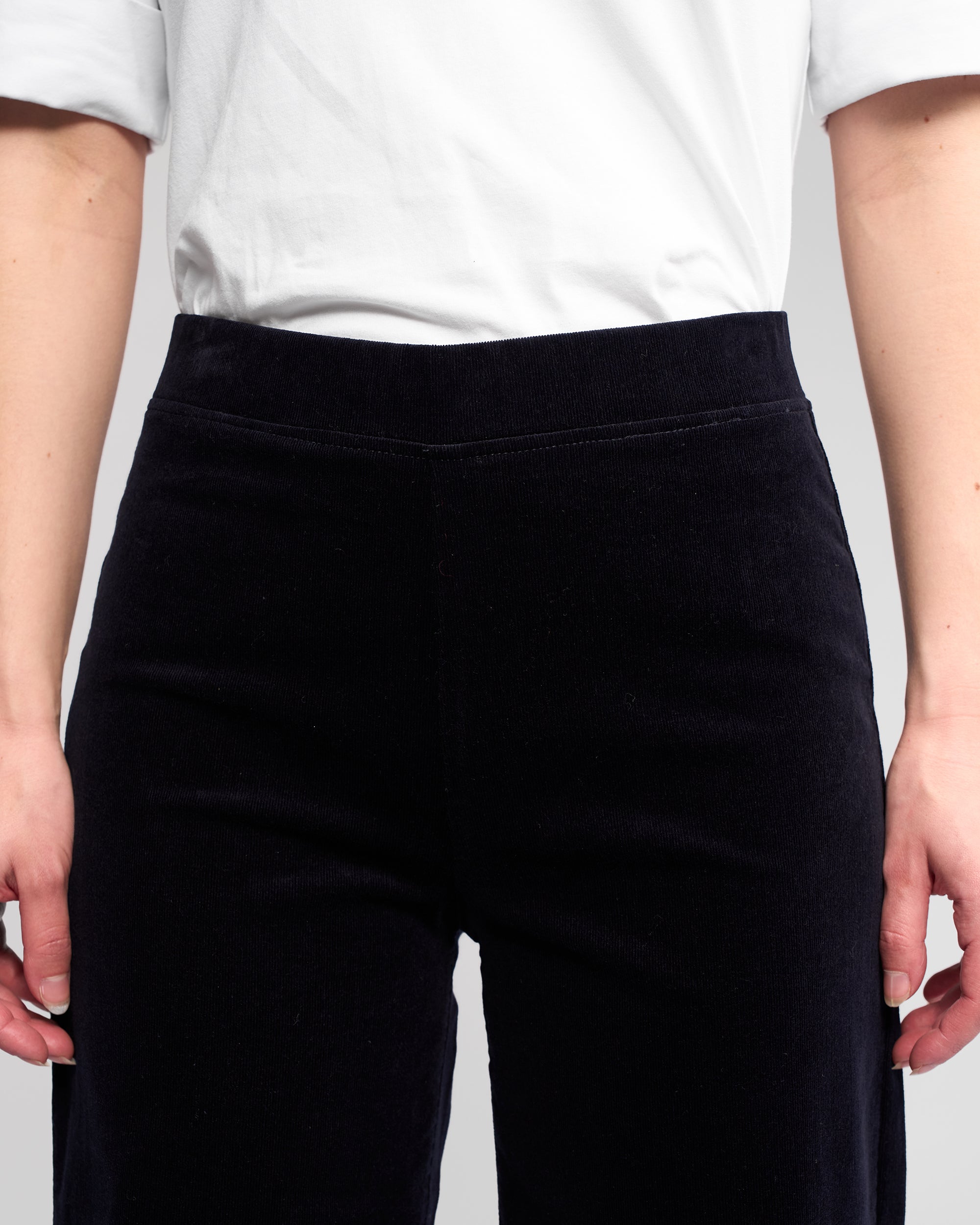 A person wearing a white short-sleeve shirt and Vassalli Loft Cord Pull On - Midnight high-waisted, wide-leg pants, shown from chest to upper thighs with arms relaxed at their sides.