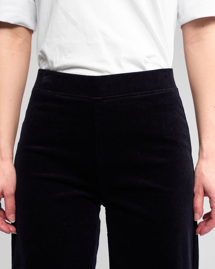 A person wearing a white short-sleeve shirt and Vassalli Loft Cord Pull On - Midnight high-waisted, wide-leg pants, shown from chest to upper thighs with arms relaxed at their sides.