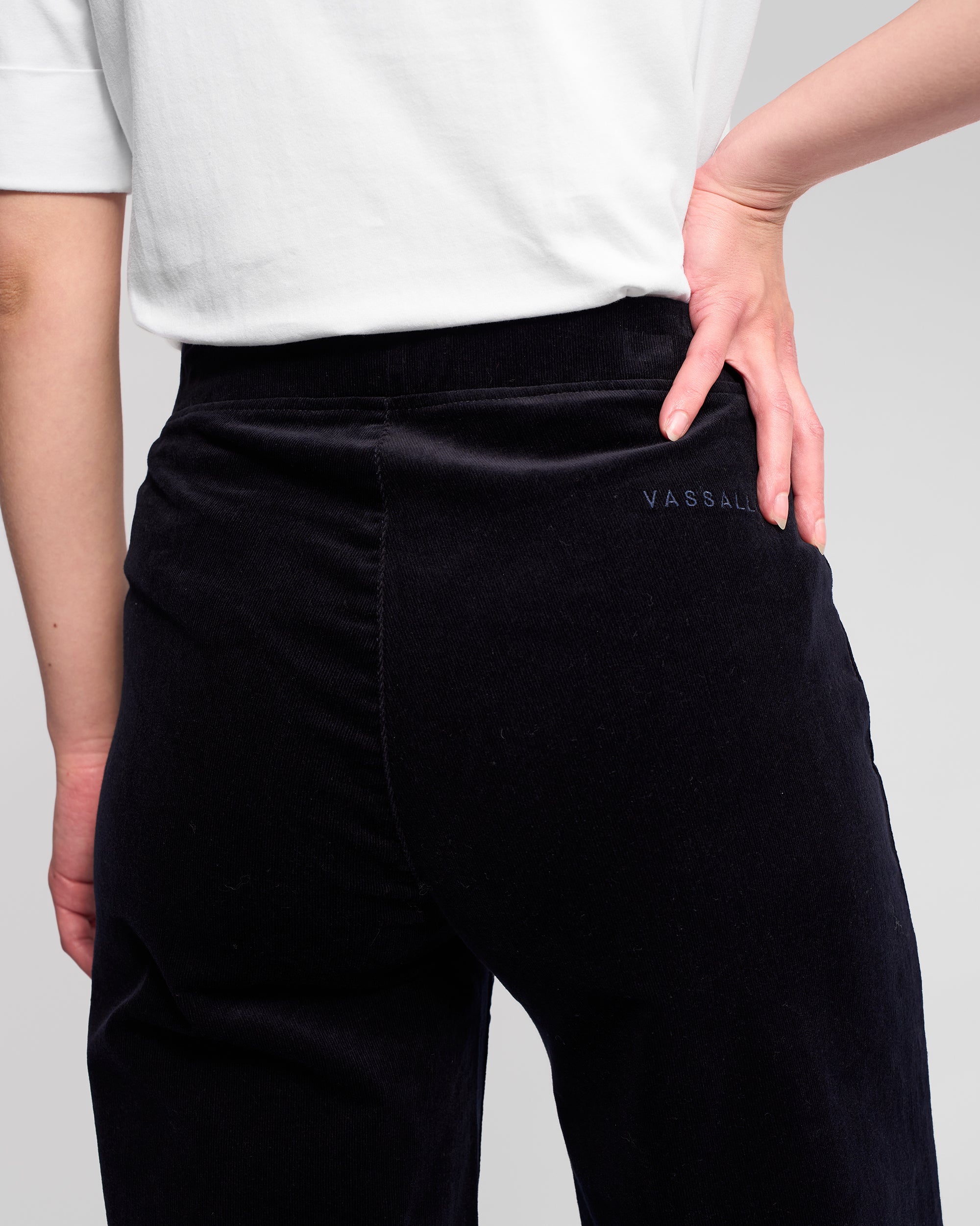 A person is shown from the back wearing a white shirt and Vassalli Loft Cord Pull On - Midnight pants, with one hand on their hip. The brand name Vassalli is embroidered on the side of the wide-leg pants.