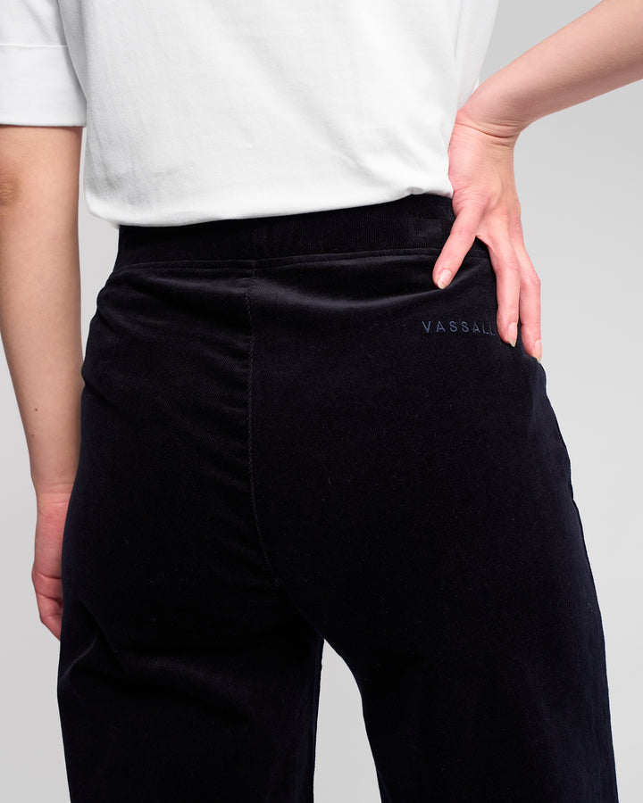 A person is shown from the back wearing a white shirt and Vassalli Loft Cord Pull On - Midnight pants, with one hand on their hip. The brand name Vassalli is embroidered on the side of the wide-leg pants.