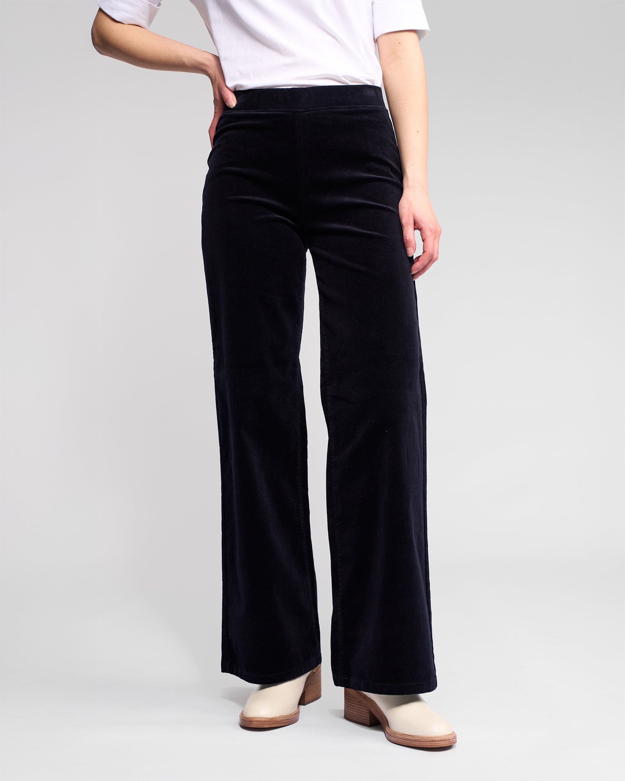 A person stands against a plain light background, wearing the Vassalli Loft Cord Pull On - Midnight high-waisted black wide-leg pants with a white shirt and cream heeled shoes. The upper body is partially out of frame.