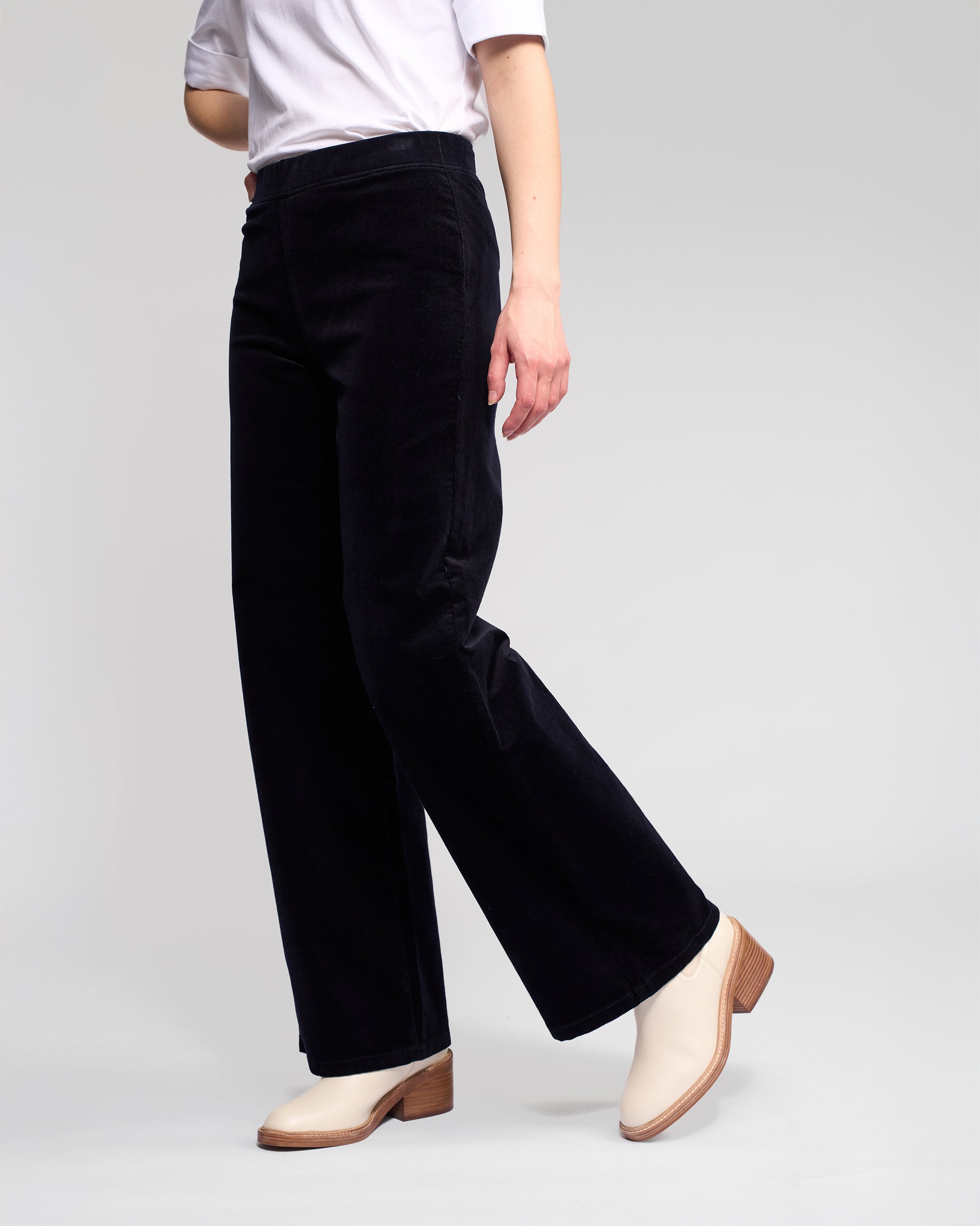 A person models the Vassalli Loft Cord Pull On - Midnight pants with a white short-sleeve top and cream heeled boots against a plain light gray background. The upper head and left hand are not visible in the frame.