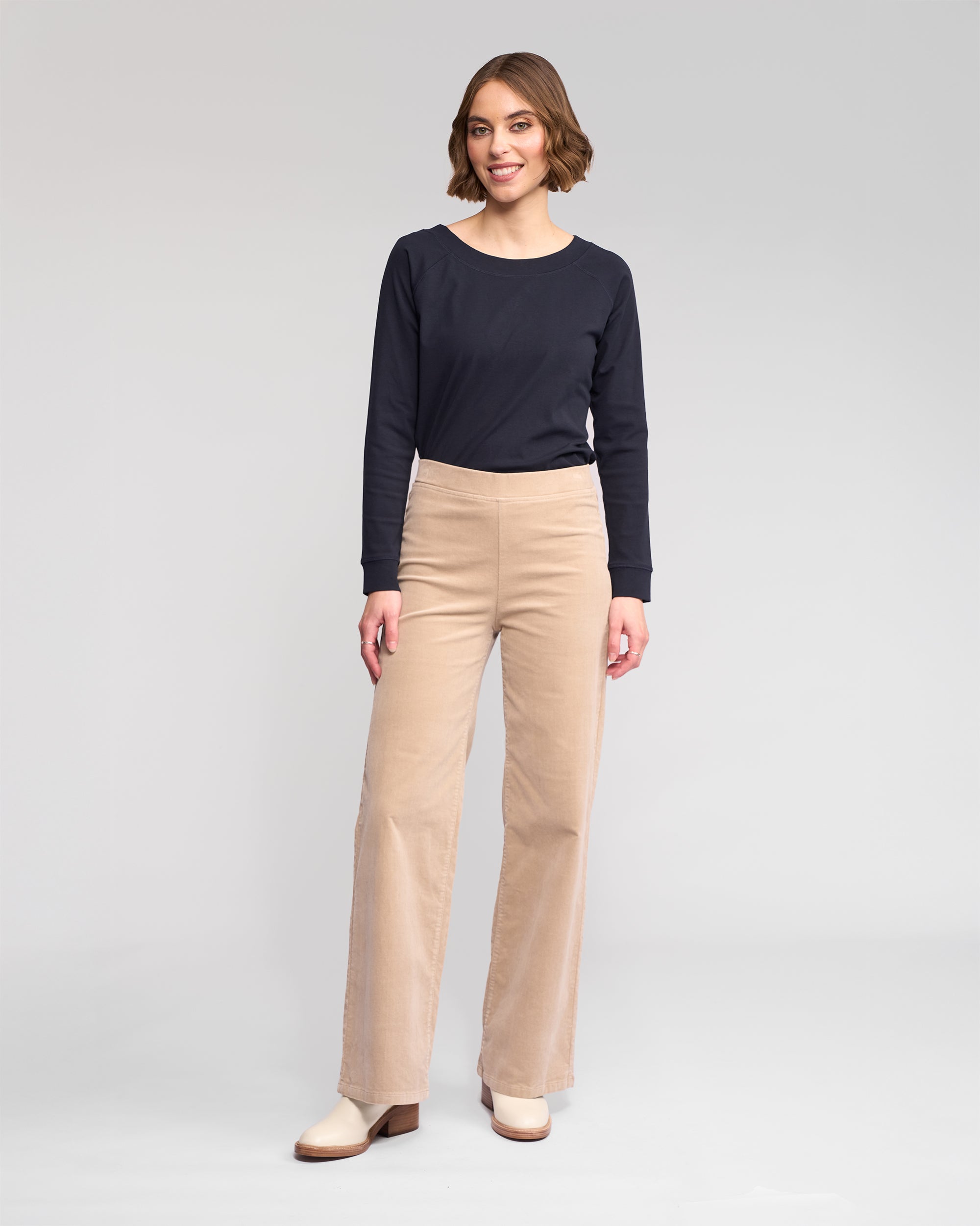 A woman with short brown hair stands smiling, wearing a long-sleeve black top, Vassalli Loft Cord Pull On pants in Oat, and cream shoes against a plain light gray background.