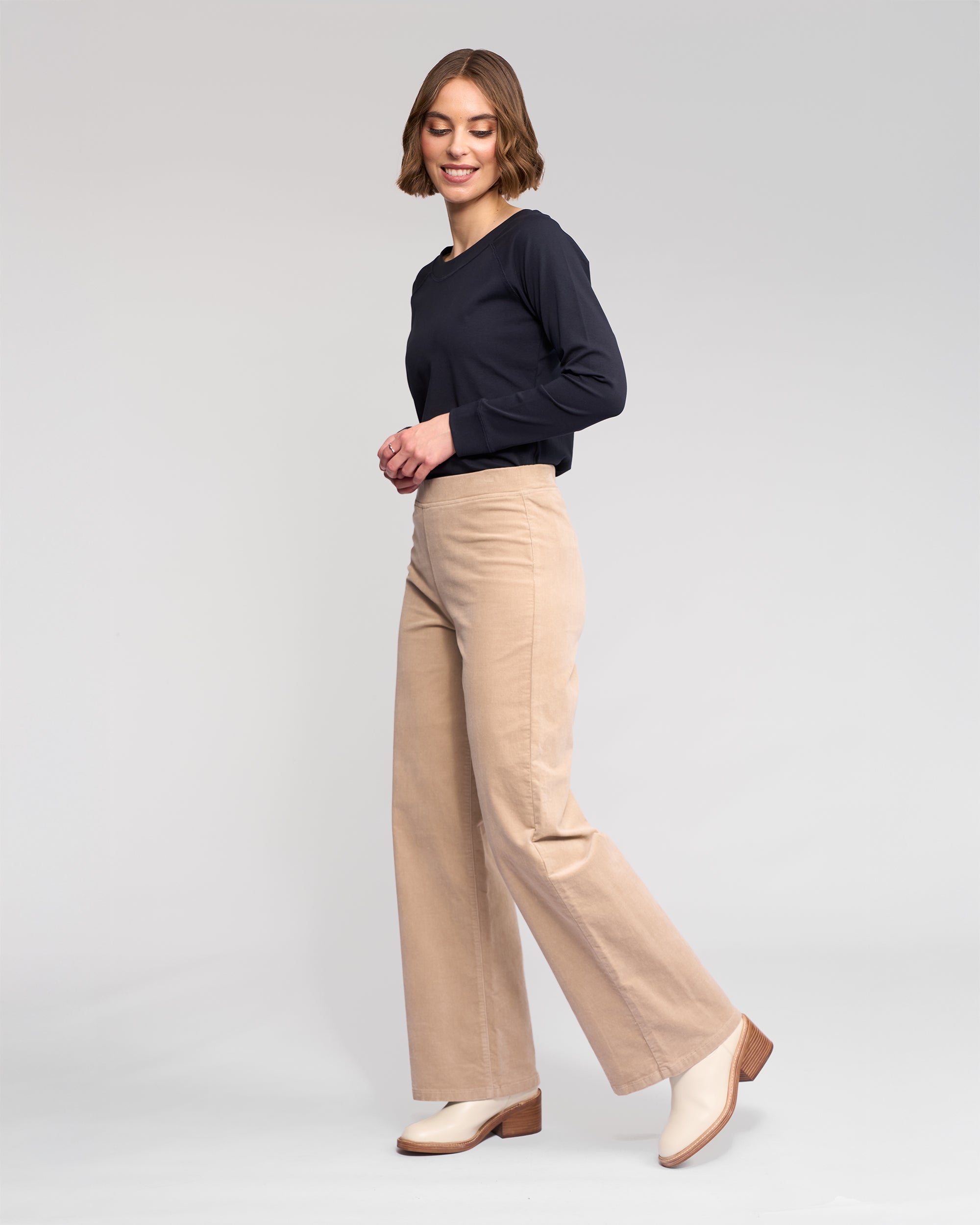 A woman with short brown hair wears a long-sleeve black top and Vassalli Loft Cord Pull On - Oat wide-leg pants, paired with cream heeled boots, standing sideways and smiling against a plain light gray background.