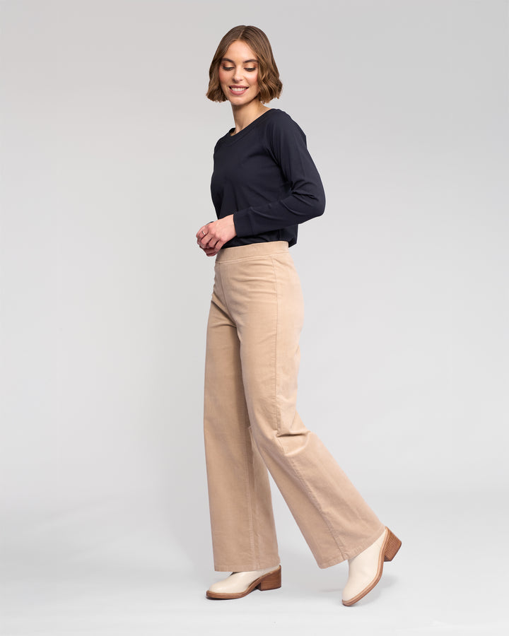A woman with short brown hair wears a long-sleeve black top and Vassalli Loft Cord Pull On - Oat wide-leg pants, paired with cream heeled boots, standing sideways and smiling against a plain light gray background.