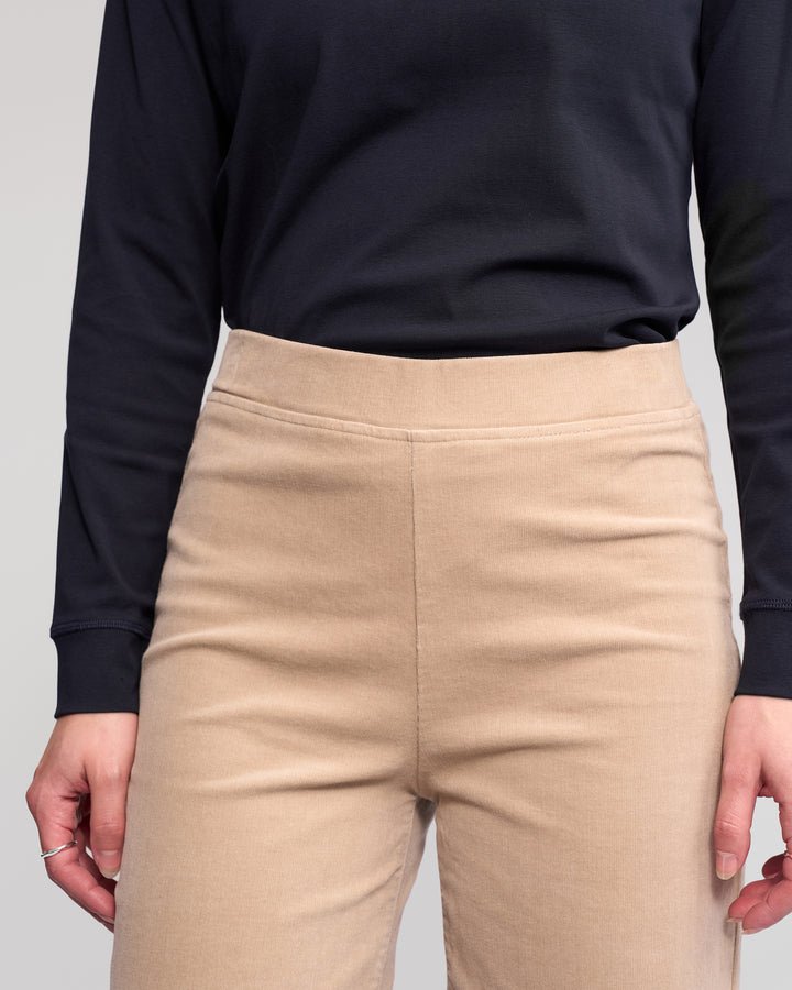 A person is shown from shoulders to thighs, wearing a long-sleeve black top with Vassalli's Loft Cord Pull On - Oat high-waisted stretch pinwale cord wide-leg pants, hands relaxed at their sides.