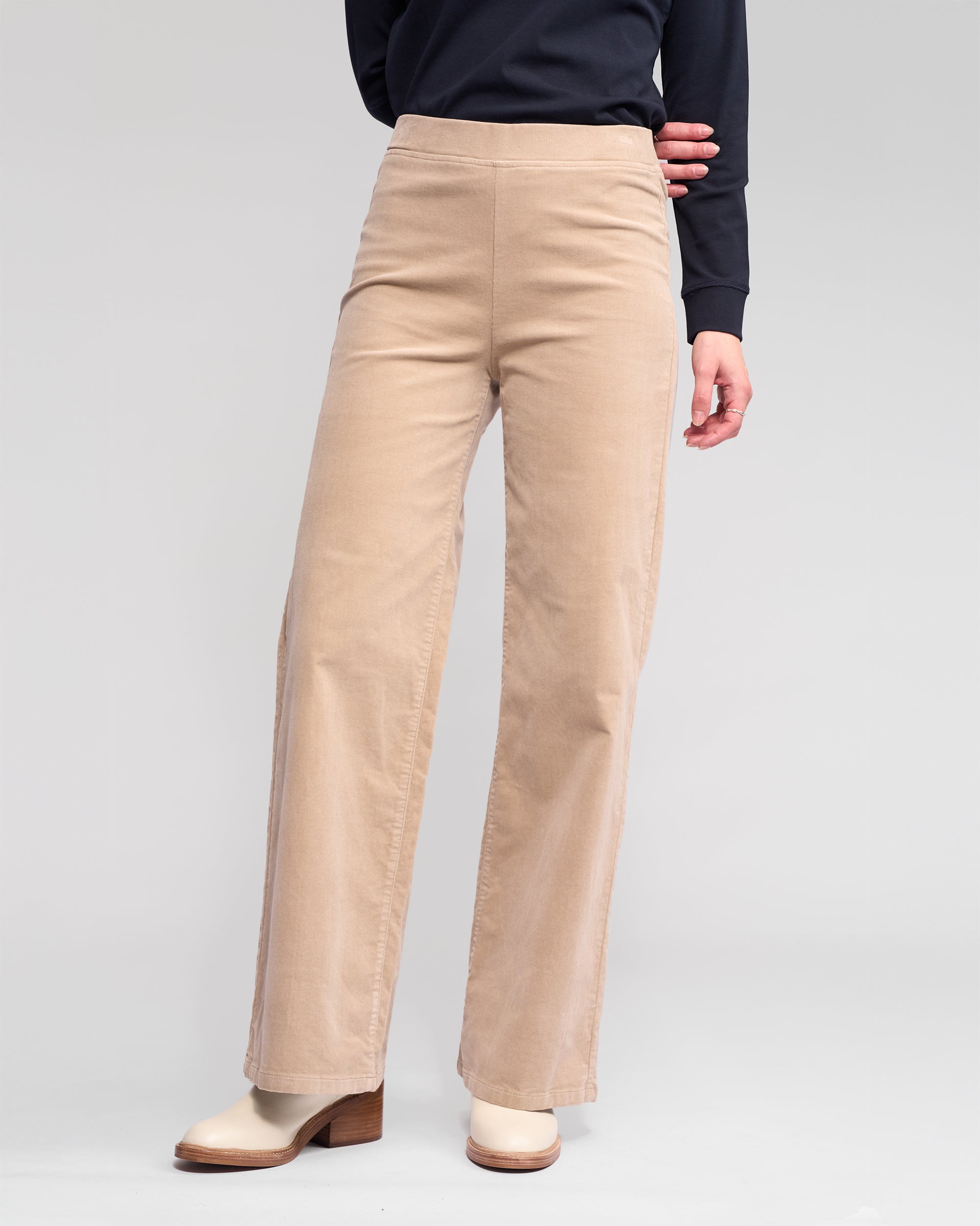 A person wearing Vassalli's Loft Cord Pull On - Oat pants, a long-sleeve black top, and off-white heeled boots stands against a plain light gray background.