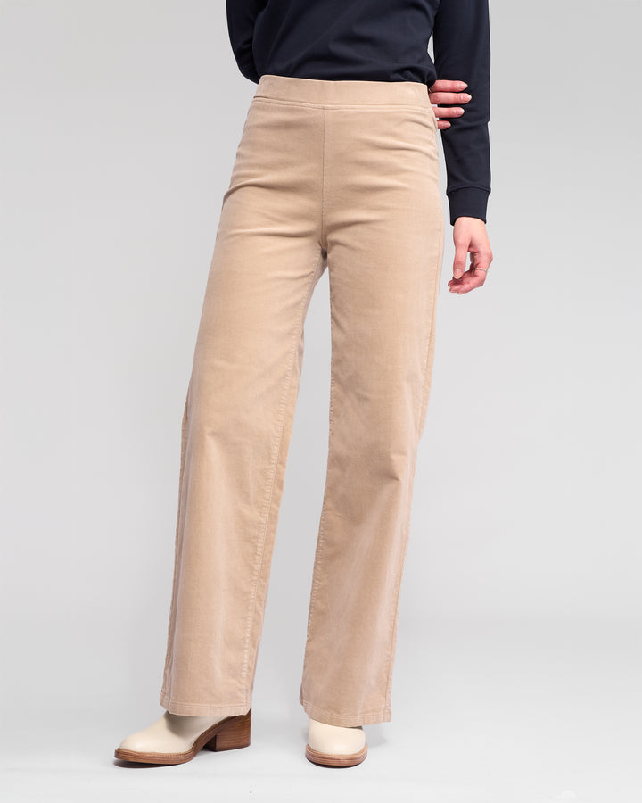 A person wearing Vassalli's Loft Cord Pull On - Oat pants, a long-sleeve black top, and off-white heeled boots stands against a plain light gray background.