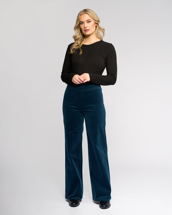 A woman with long blonde hair stands against a plain light gray background, wearing the Vassalli Loft Cord Pull On - Petrol wide-leg pants, a black long-sleeve shirt, and black shoes. She faces forward with her hands gently clasped.