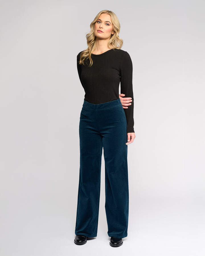 A woman with long, wavy blonde hair stands against a plain background, wearing black shoes and Vassalli’s Loft Cord Pull On - Petrol high-waisted wide-leg pants with a fitted long-sleeve black top.
