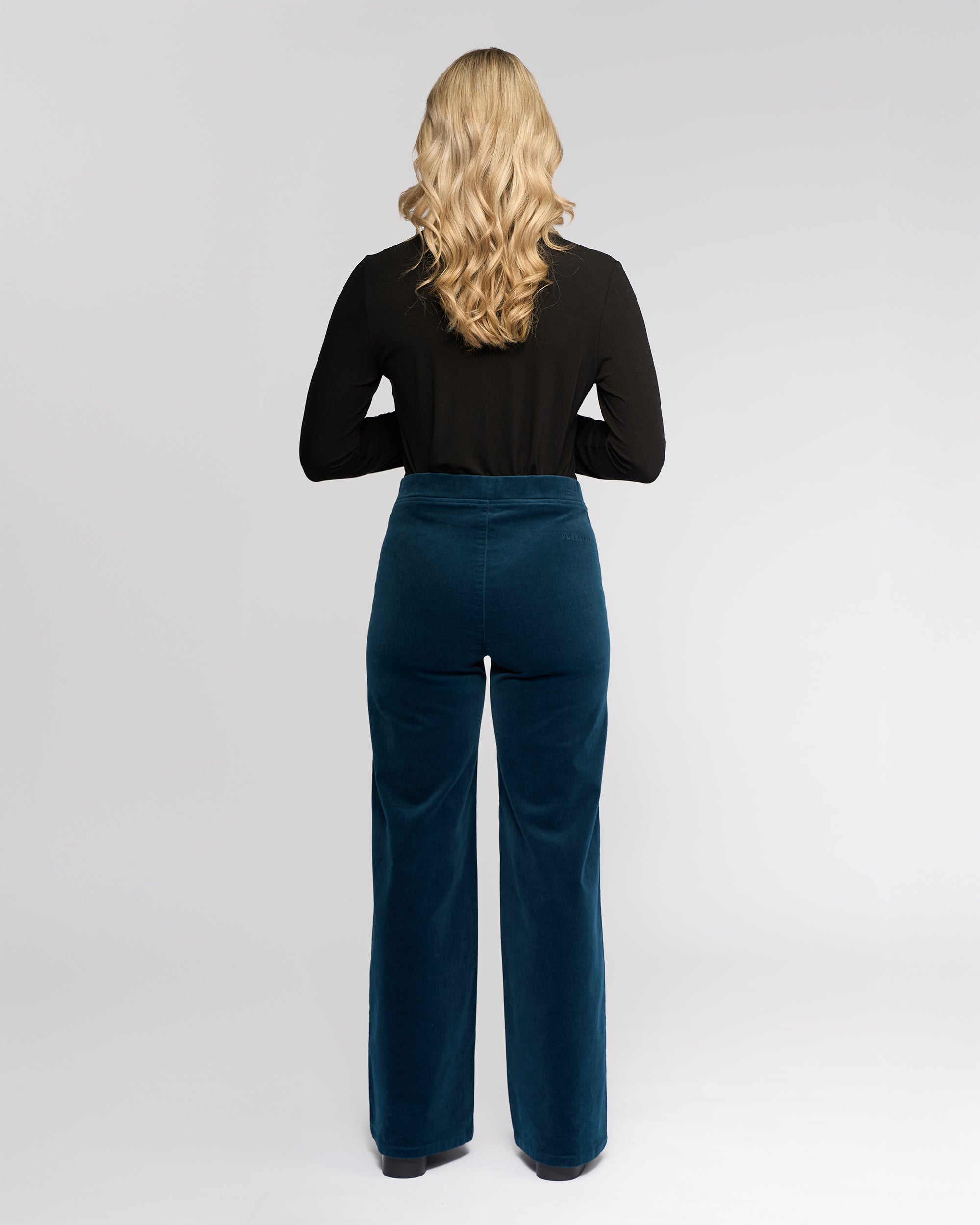 A woman with long, wavy blonde hair stands facing away, wearing a black long-sleeve top and Vassalli's Loft Cord Pull On - Petrol pants against a plain light background.