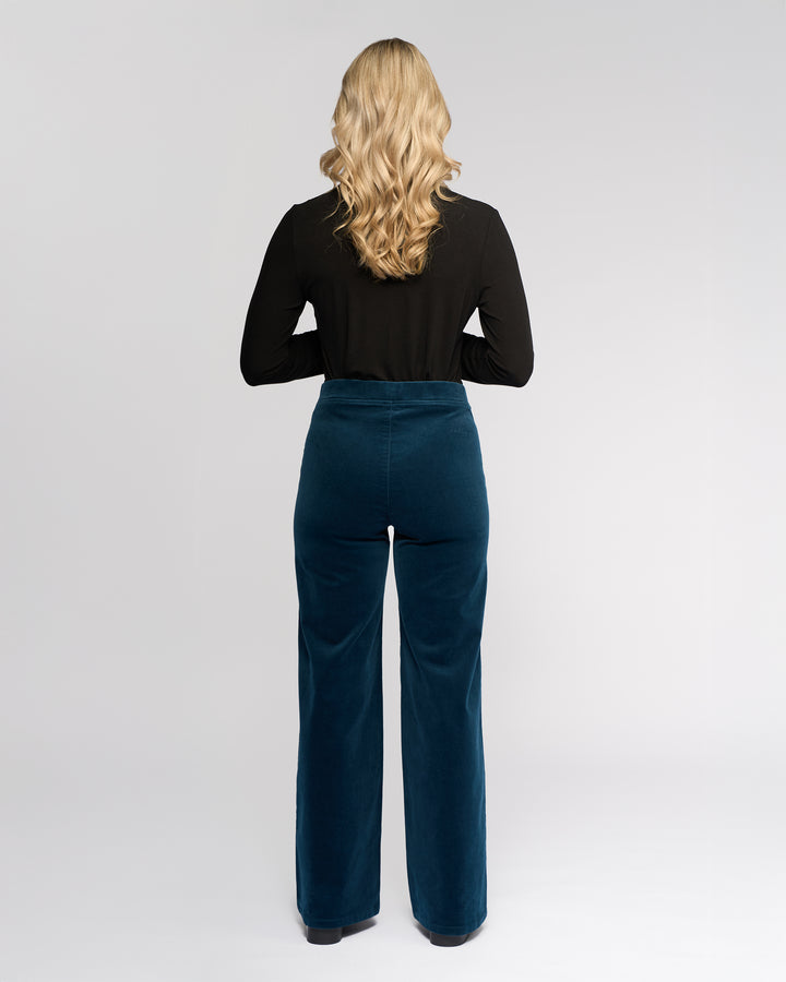 A woman with long, wavy blonde hair stands facing away, wearing a black long-sleeve top and Vassalli's Loft Cord Pull On - Petrol pants against a plain light background.